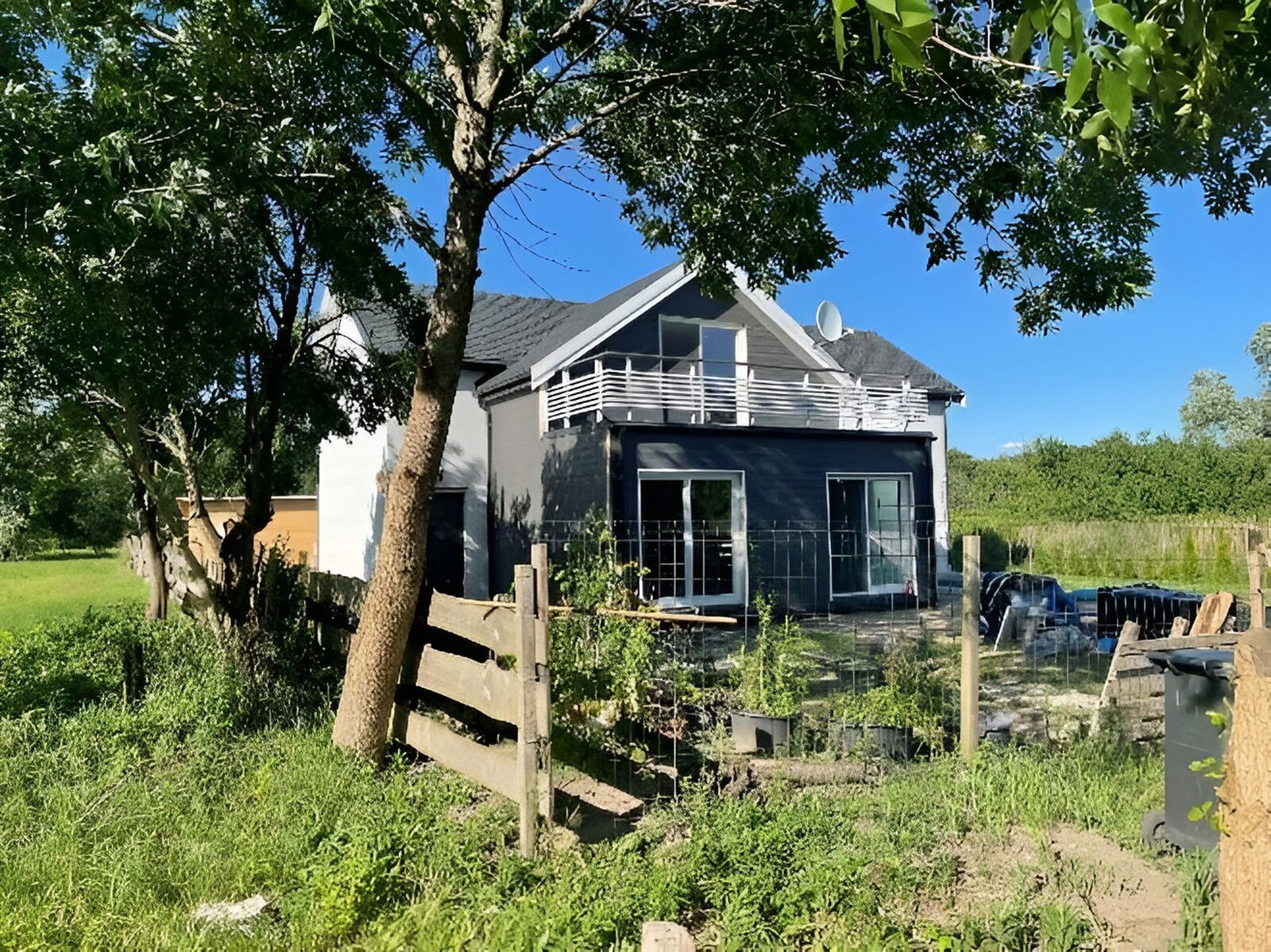 Fotografia przedstawia mały, domek letniskowy ogrodzony drewnianym płotem, wokół zarośla. 