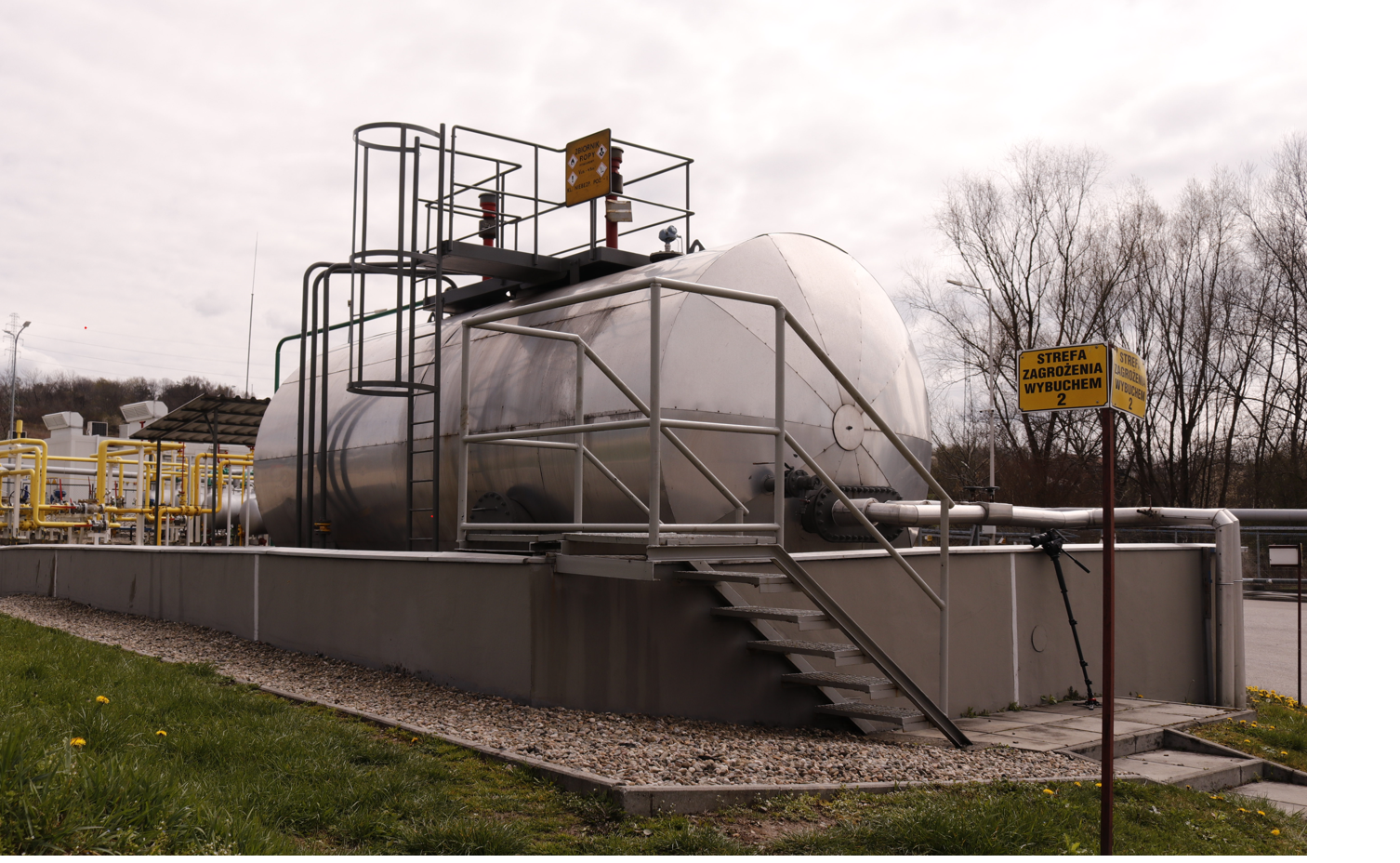 Zdjęcie przedstawia zbiornik magazynowy na ropę z wejściem oraz tablicami informacyjnymi. Zbiornik ma cylindryczny kształt. Jest zorientowany poziomo i osadzony na platformie. Prowadzą do niej schody. Natomiast do górnej części zbiornika poprowadzona jest drabinka. Na górze znajduje się nieduży podest, umożliwiający dostęp do zbiornika obsłudze. Na pierwszym planie, po prawej stronie znajduje się żółta tablica informacyjna o następującej treści: strefa zagrożenia wybuchem - druga. W tle po prawej stronie znajdują się drzewa. A po lewej infrastruktura napowierzchniowa, umożliwiająca transport i magazynowanie ropy.