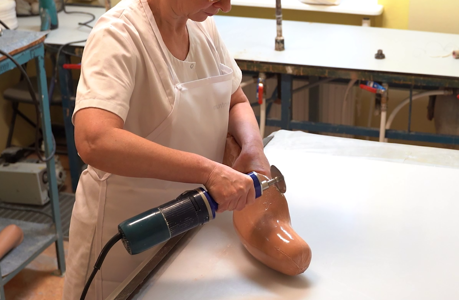 Zdjęcie przedstawia kobietę pracującą przy stole z piłą oscylacyjną. Urządzenie trzyma w prawej dłoni. Kobieta ma na sobie biały strój roboczy. Piła oscylacyjna wyposażona jest w przewód, walcowatą część z uchwytem oraz w tarczę. Tarcza stanowi element umożliwiający manipulację laminatem czy też gipsem. Jest ona zamocowana na wale znajdującym się w osi podłużnej urządzenia. W drugiej ręce kobieta trzyma fragment protezy, który opracowuje. Za nią znajduje się stół do laminacji.