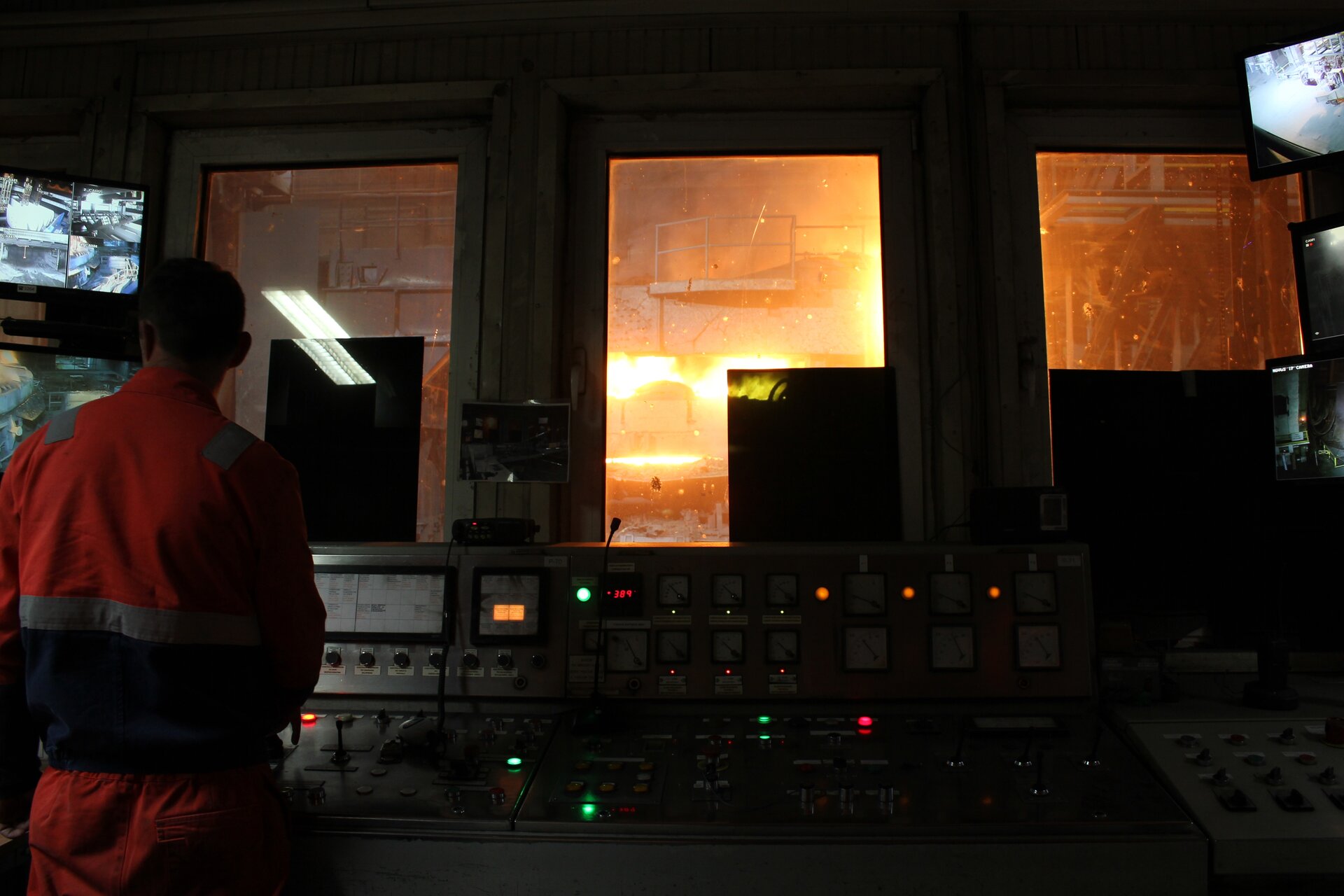 Zdjęcia przedstawia operatora monitorującego przebieg procesu w elektrycznym piecu łukowym. . Na pierwszym planie widoczny jest pulpit sterowniczy. Znajduje się na nim duża ilość przycisków, dźwigni oraz tarcz manometrów. Dookoła widoczne są monitory. Z lewej strony pulpitu widoczny jest operator spoglądający na monitory po lewej stronie. Operator ubrany jest w pomarańczowy strój roboczy. Na drugim planie widoczne są trzy okna. Przez okna widać metalową konstrukcję, balkony, schody oraz iskrę.