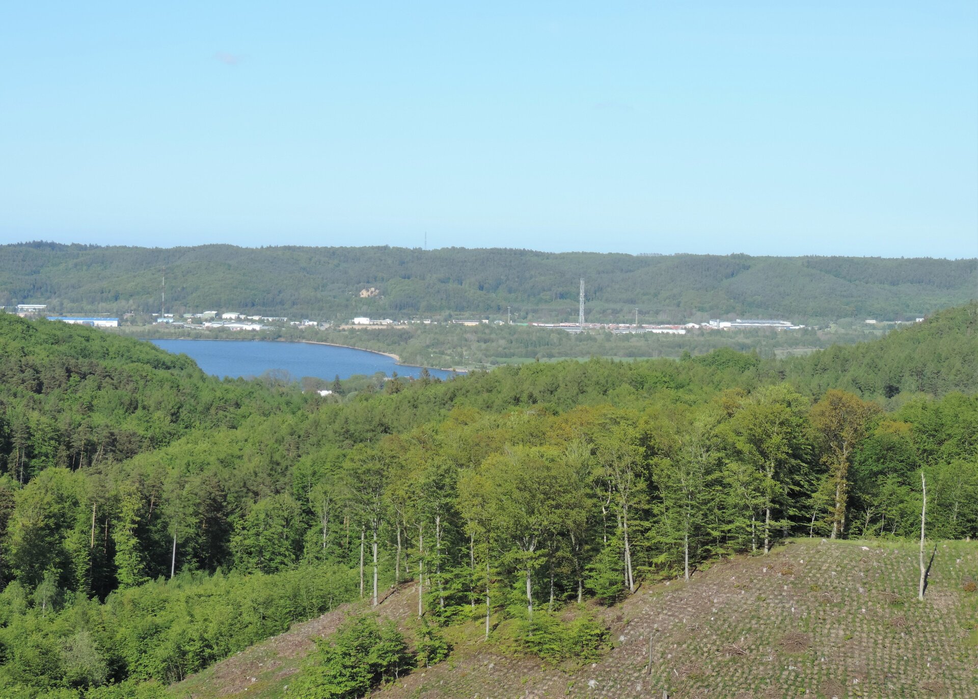 Zdjęcie przedstawia widok ze wzgórza na jezioro otoczone lasem znajdujące się na drugim planie. Na pierwszym planie zalesione wzniesienie.