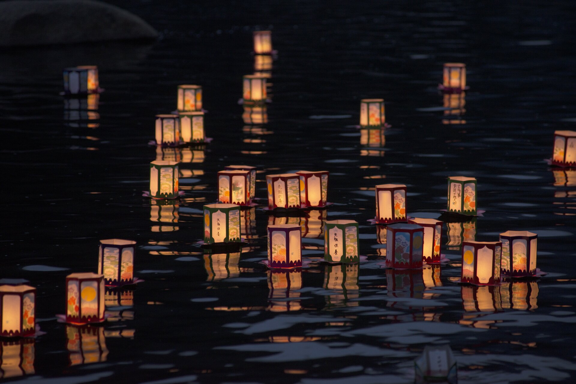 Ilustracja przedstawiająca lampiony pożegnalne, które pływają na wodzie. Lampiony są białe z brązowymi obwódkami. Z lampionów wydobywa się światło.