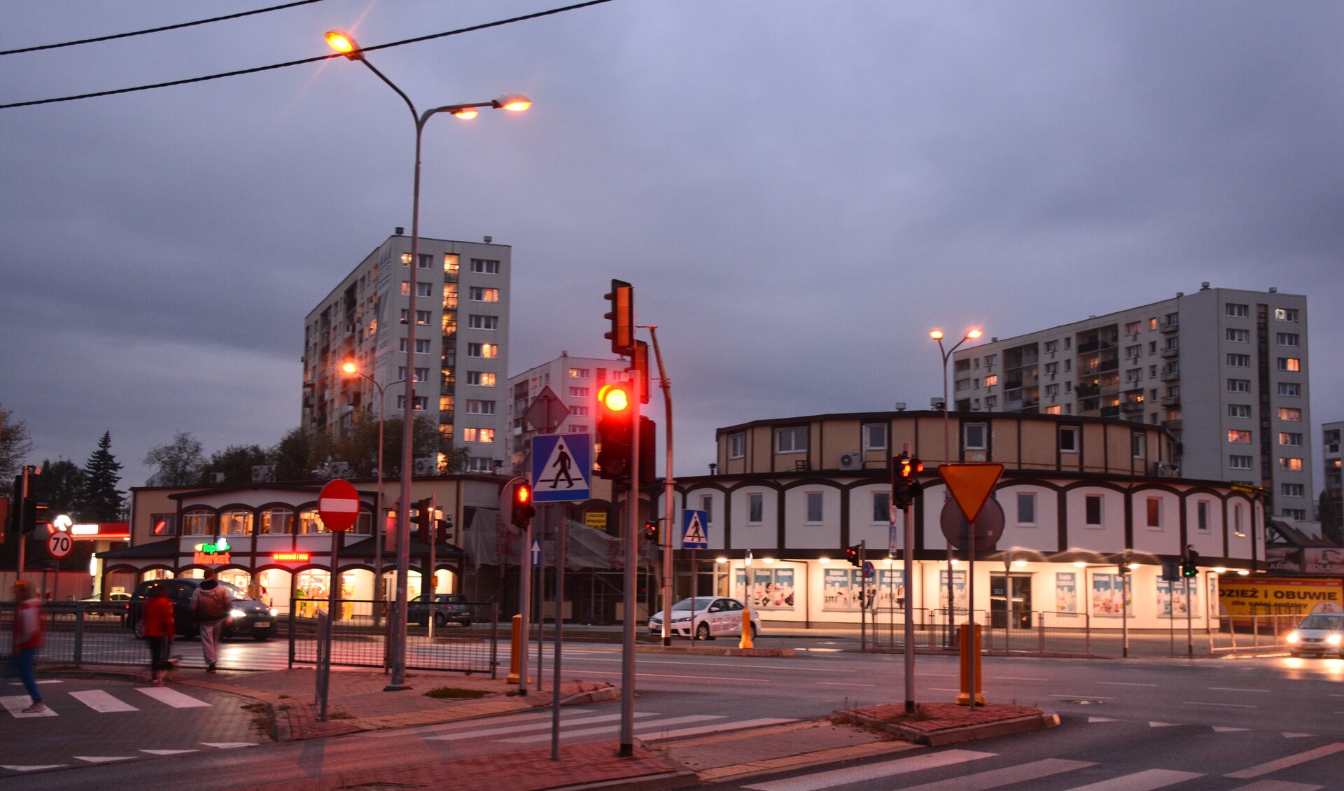Zdjęcie przedstawia Legionowo o zmierzchu. Na pierwszym planie jest skrzyżowanie. Na drugim planie jest pasaż handlowy i bloki.