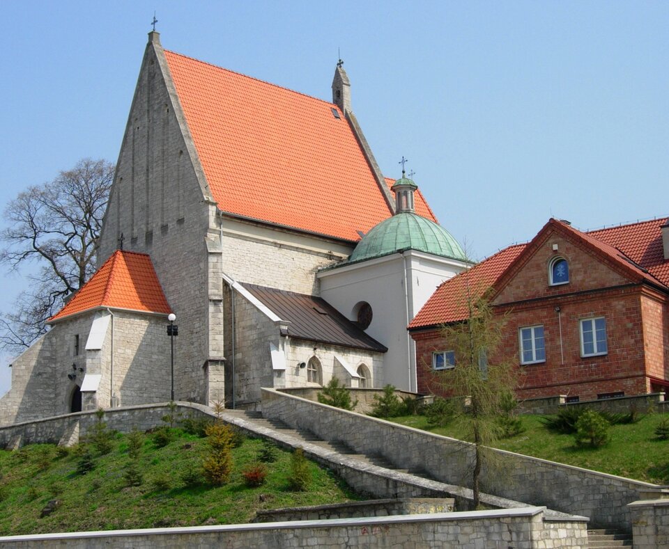 Fotografia przedstawia budynek zbudowany z kamienia ze spadzistym trójkątnym dachem, na czubku którego jest krzyż. Obok niska przybudówka z okrągłym dachem, niską wieżą i krzyżem. Budynek stoi na wzniesieniu i prowadzą do niego schody. Obok widać ceglany budynek mieszkalny.