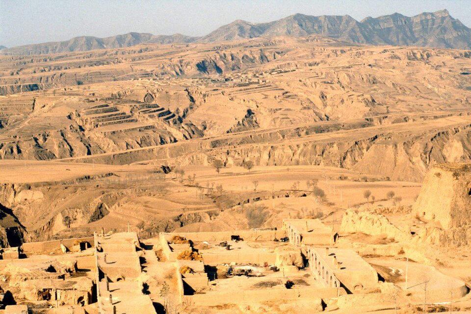 Na zdjęciu Wyżyna Lessowa. Dominuje kolorystyka żółtopomarańczowa. Na pierwszym planie zabudowania w kolorze podobnym do koloru lessu. Dalej różne formy lessowe, zagłębienia, doliny, tarasy. W tle góry. Na horyzoncie niebo.