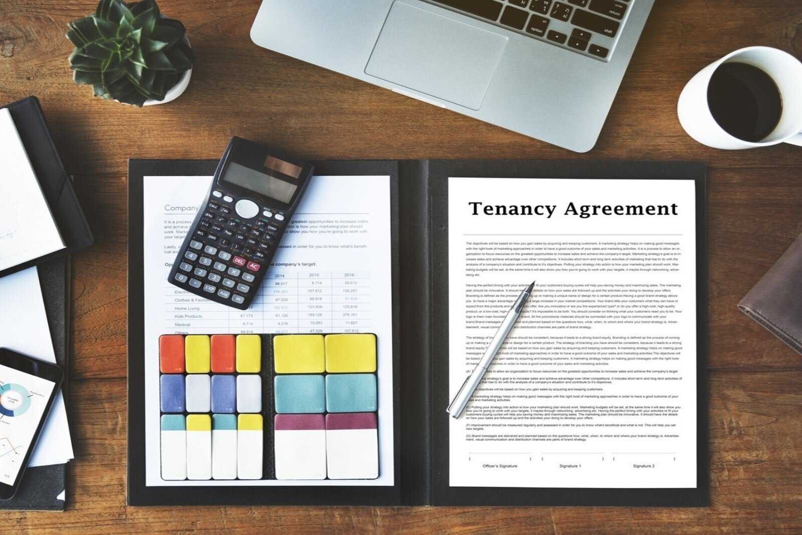 Zdjęcie przedstawia biurko w ujęciu z góry na którym leżą: teczka z dokumentem umowy najmu. Na dokumencie leży kalkulator, znaczniki stron w różnych kolorach i długopis. Nad teczką widać fragment otwartego laptopa, kwiat doniczkowy oraz kubek z kawą.