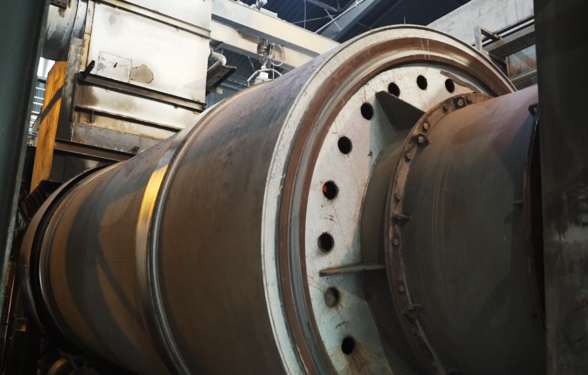 Zdjęcie przedstawia dużą, stalową konstrukcję pieca obrotowego. Złożona jest ona z dużego walca ułożonego w poziomie, połączonego rurami z innymi częściami pieca.