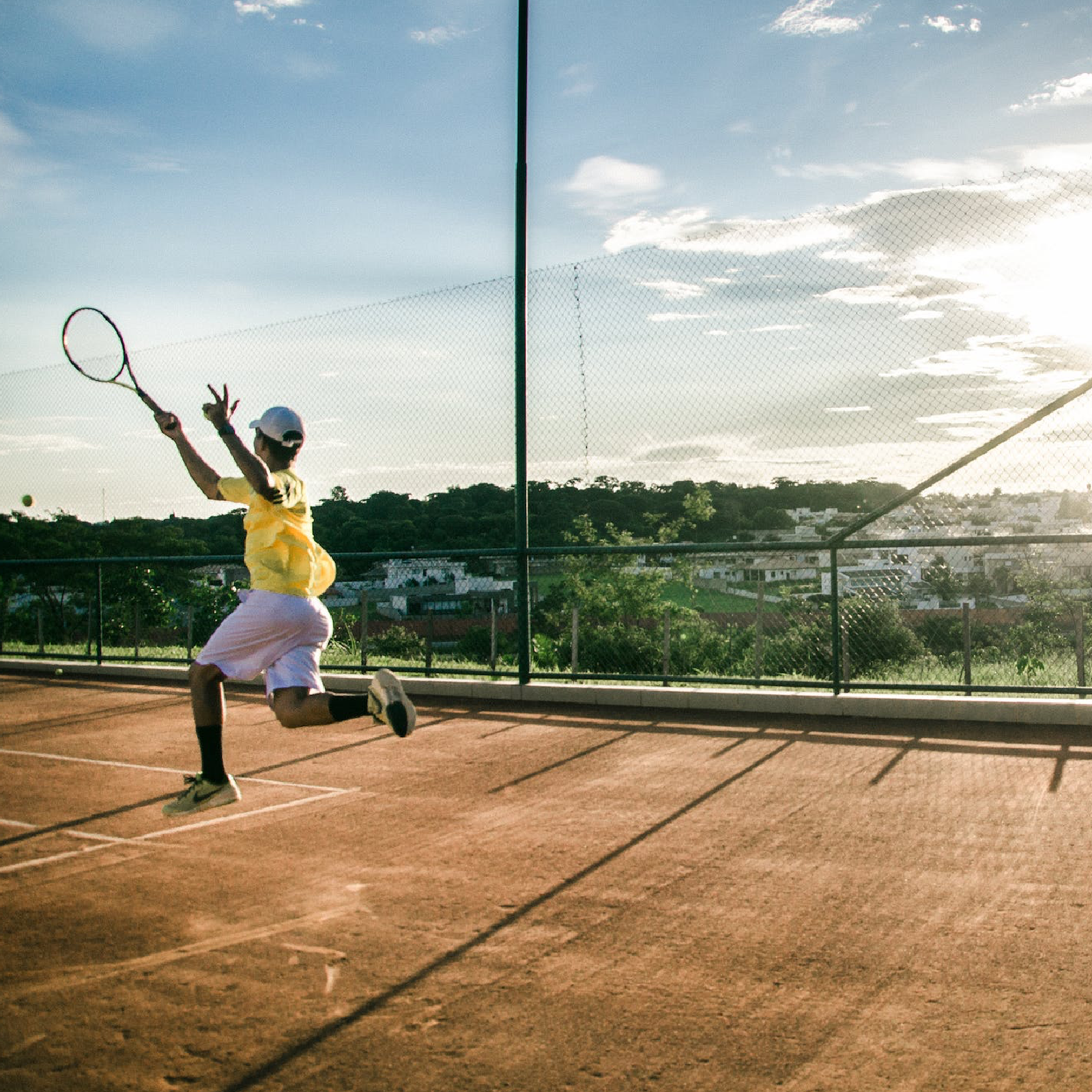 Ilustracja przedstawia kobietę biegnącą z rakietą na drugi koniec kortu do tenisowego. Podłoże wykonane jest z ubitego, pomarańczowego piasku. W tle widoczne są budynki. Kort otoczony jest siatką.