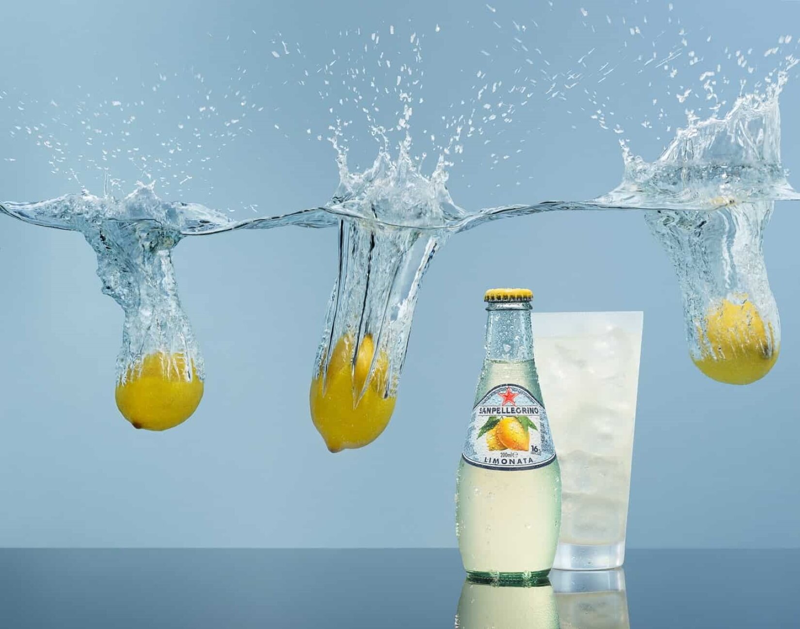 Fotografia przedstawia naczynie z wodą. Nie widać jego krawędzi. Poniżej powierzchni wody ustawiono szklaną butelkę z napojem. Napój jest w jasnym, żółtawym kolorze, a za nim stoi szklanka wypełniona lodem i płynem o podobnym odcieniu. Na etykiecie umieszczonej na butelce widoczne są dwie żółte cytryny z zielonymi listkami, a także napis „LIMONATA”, oraz logo – czerwona gwiazdka. Butelka posiada żółty kapsel. W okolicy wysokości jej szyjki znajdują się trzy cytryny, wrzucone do wody. Dwie są z lewej strony butelki, a jedna – z prawej. Cytryny wzburzyły powierzchnię wody, wyrzucając do góry jej krople, a także zostawiając ślad w jej powierzchni. Woda jak gdyby zatrzymała się w miejscu, w którym jej powierzchnię naruszyły cytryny, dokładnie pokazując ich ślady.
