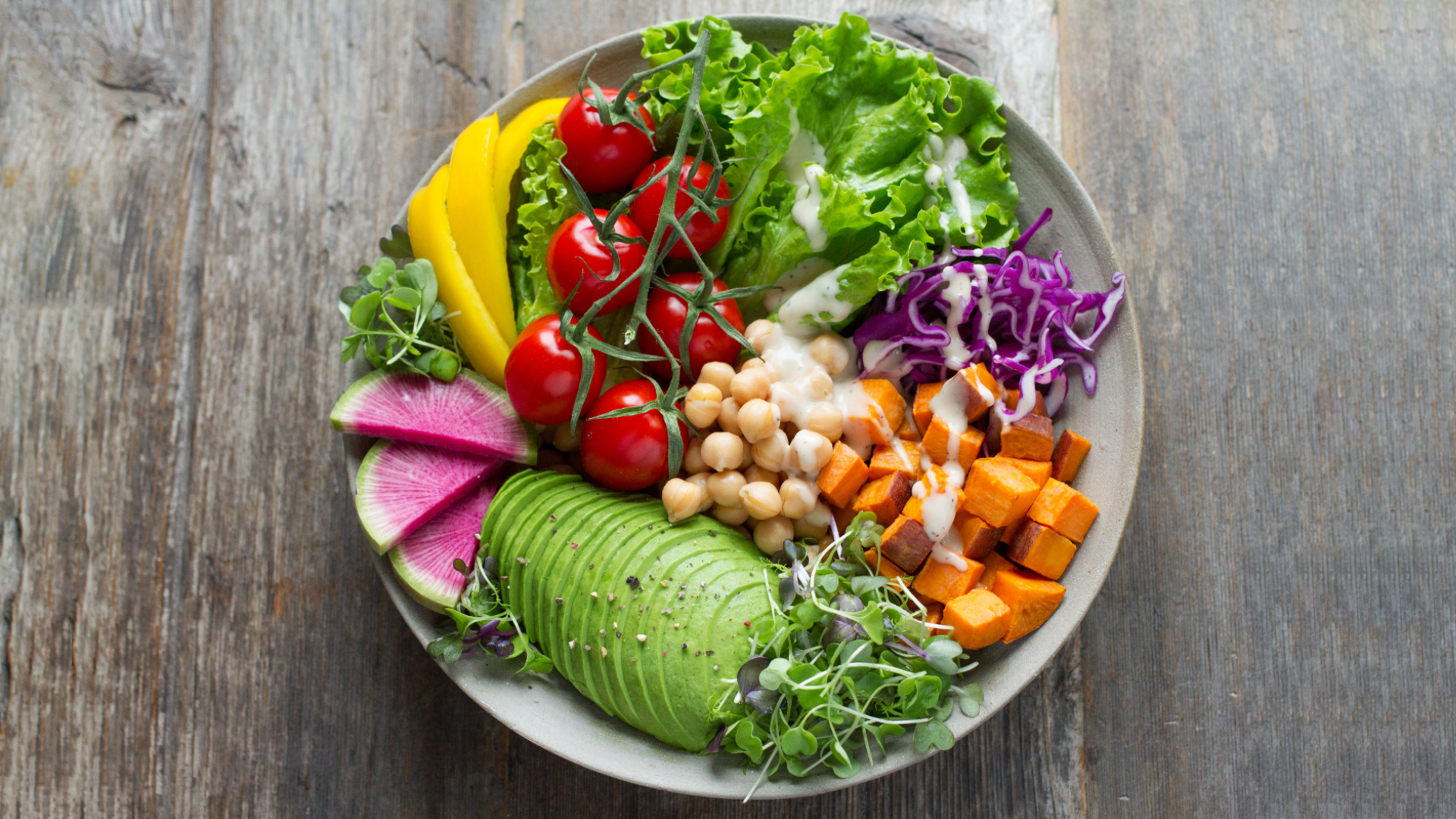 Zdjęcie wykonane z góry przedstawia talerz pełen warzyw stojący na drewnianym stole z desek. Widoczne są pomidorki koktajlowe na gałązce, liście sałaty, poszatkowana czerwona kapusta, dynia w kostkach, pokrojone w plastry awokado, kilka plasterków rzodkwi arbuzowej. Pomiędzy warzywami ułożone są różnego rodzaju kiełki. Po środku leży ciecierzyca.
