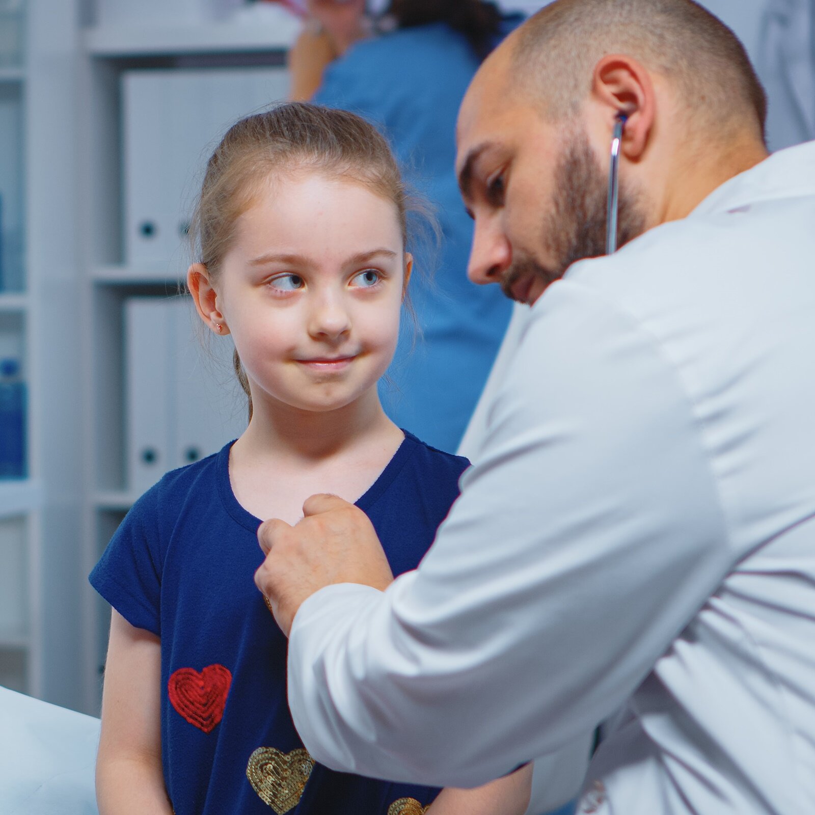 Zdjęcie przedstawia mężczyznę, w ujęciu od pasa w górę, w białym fartuchu lekarskim i z włożonymi do uszu słuchawkami stetoskopu. Mężczyzna widoczny jest z lewego boku i zwrócony w kierunku dziewczynki w wieku szkolnym. Słuchawkę stetoskopu przykłada do klatki piersiowej dziewczynki, która, z lekkim uśmiechem, spogląda na jego twarz. 