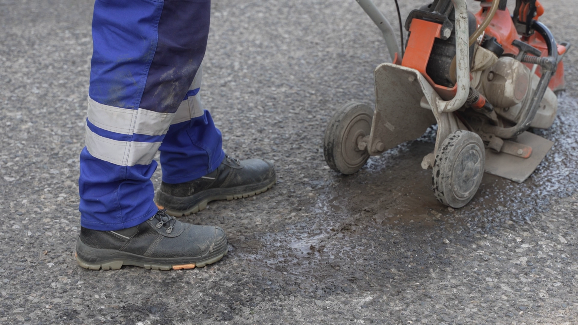 Na grafice przedstawiono masywne buty ochronne ze wzmocnieniami. Osoba nosząca je idzie po asfalcie i pcha maszynę.
