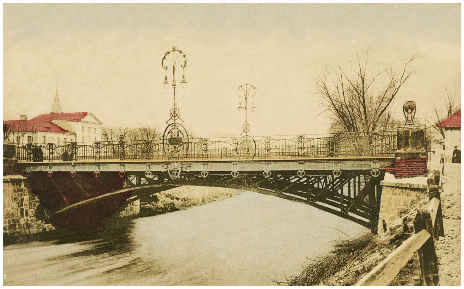 Zdjęcie w sepii przedstawiające stalowy most wiszący nad rzeką, z kratownicą pod jezdnią zbudowaną z prętów połączonych łukowymi przęsłami. Jezdnie ograniczają kute, ornamentalne barierki. No osi mostu, po obu stronach znajdują się ozdobne, kute lampy naftowe. W tle jasne budynki.