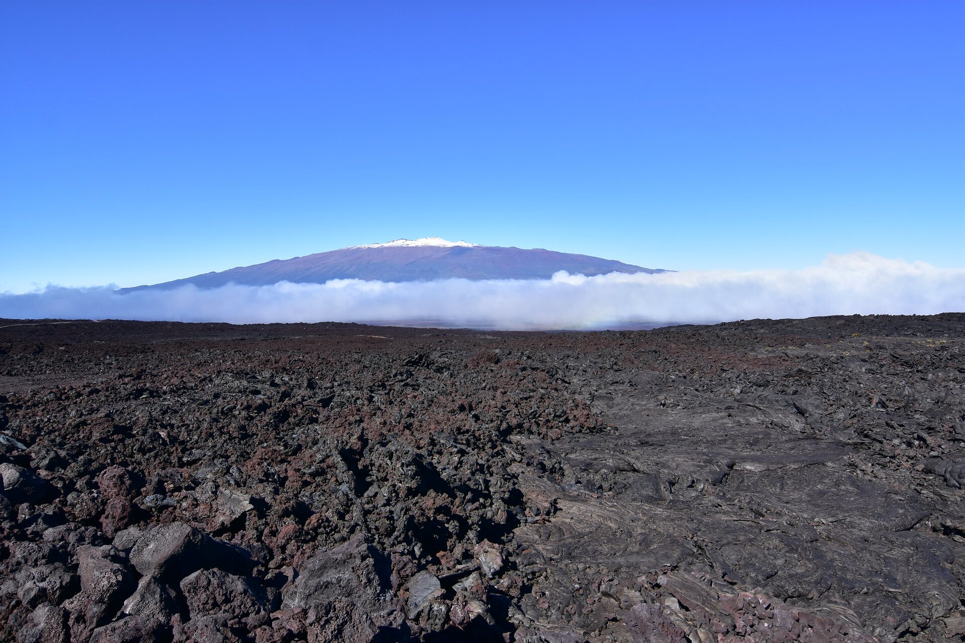 Na zdjęciu w oddali jest wulkan. Tylko szczyt jest ośnieżony. Na pierwszym planie jest płaski teren pokryty ciemnymi grudami zastygłej lawy. Niebo nad wulkanem jest bezchmurne.  