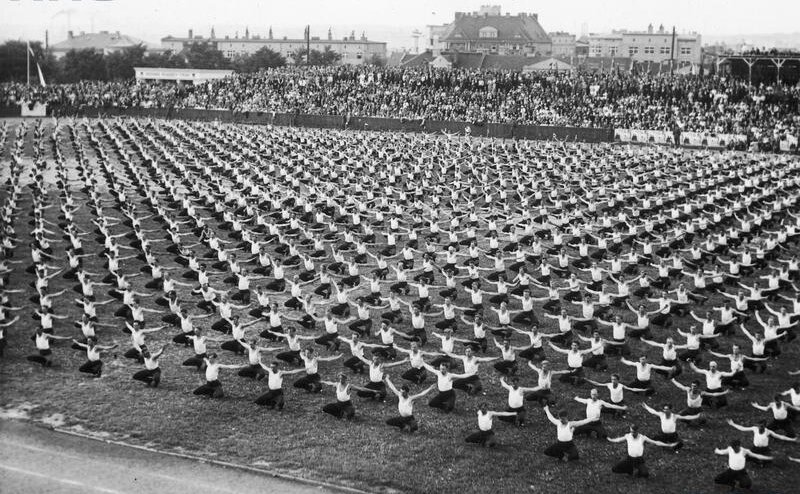 Zdjęcie przedstawia murawę, na której znajdują się sportowcy. Wszyscy mają tę samą pozę i są równomiernie rozmieszczeni. Kucają i mają szeroko rozstawione ręce. W tle widoczne są trybuny wypełnione widzami.