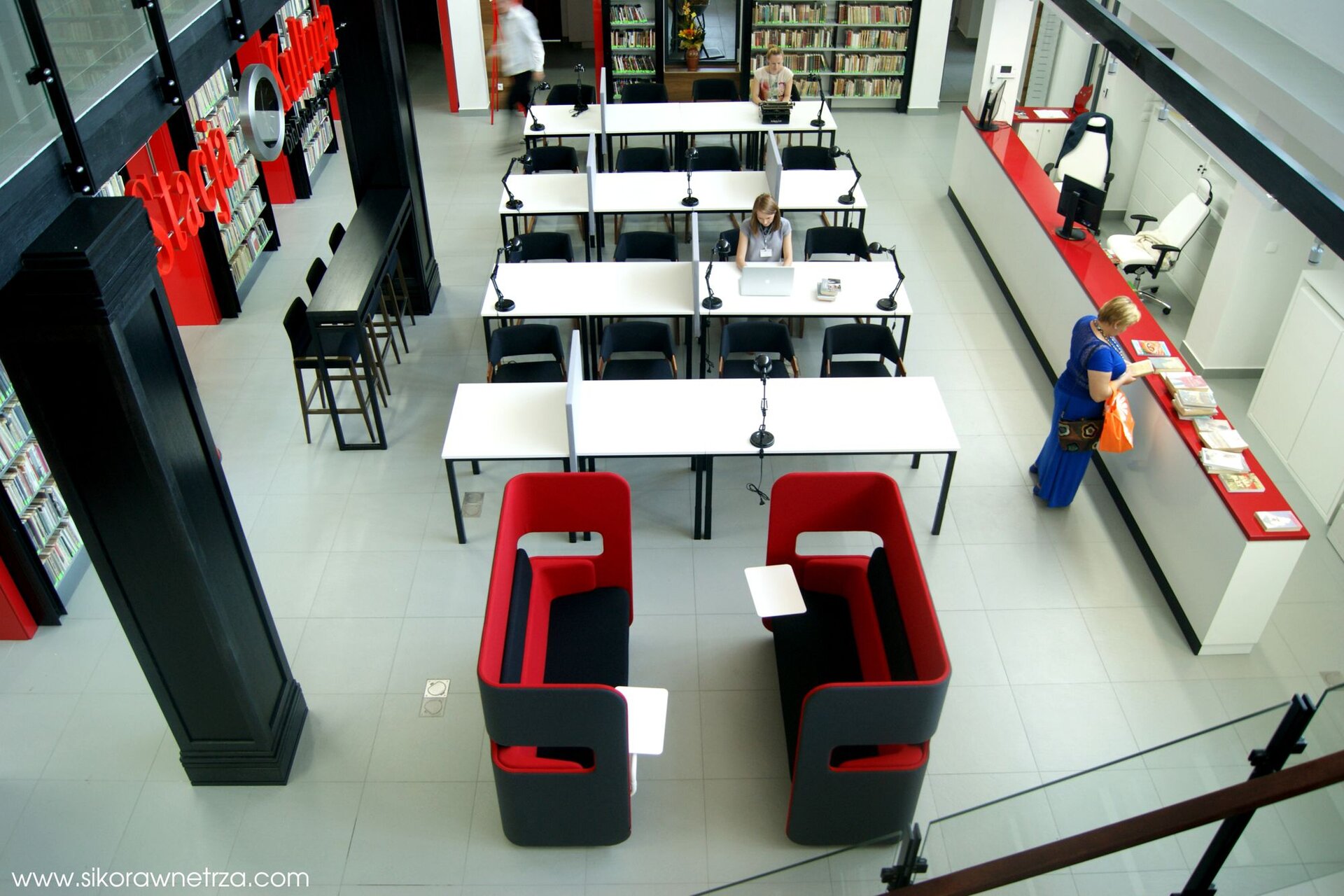 Zdjęcie przedstawia widok z góry na pomieszczenie w bibliotece. Na środku stoją stoliki czytelnicze, przy których siedzą dwie osoby. 
