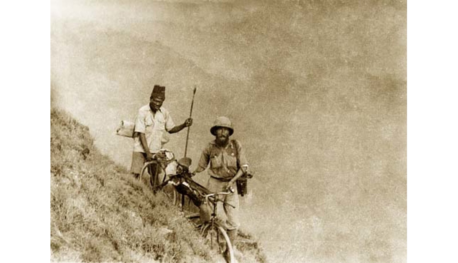Zdjęcie w sepii przedstawiające dwóch mężczyzn. Jeden z nich podpiera się na kiju, drugi prowadzi rower.