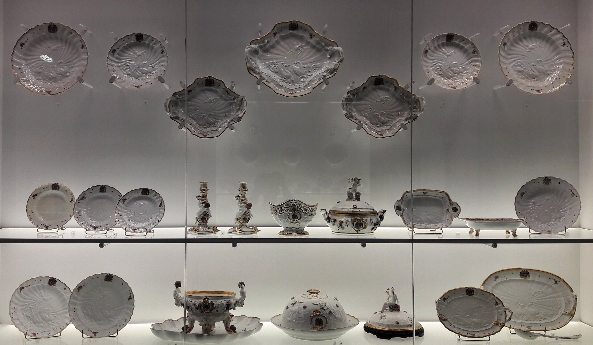 Kliknij, aby powiększyć. Zorientowana poziomo fotografia barwna ukazująca fragment gabloty, w Muzeum Narodowym w Warszawie, z zastawą stołową stanowiącą część tzw. serwisu łabędziego. Pośród eksponatów przeważają ustawione pionowo na stojakach za szybą na dwóch poziomych półkach, a górą zawieszonych na ścianie białe talerze i półmiski różnej wielkości i kształtów wyposażone w uszka. Łączy je motyw reliefu ukazującego na dnach parę łabędzi oraz stały motyw herbu na brzegu, pojedynczych drobnych elementów kwiatowych i złoceń krawędzi. Na dolnej półce od lewej: dwa talerze, otwarta waza stojąca na czterech nogach w podstawie w kształcie muszli, okrągły półmisek z pokrywą, pokrywa z nagą postacią kobiecą na szczycie, dwa półmiski. Na półce powyżej od lewej: trzy mniejsze talerze, dwa pojedyncze świeczniki z oplatającymi je postaciami, naczynie na owoce, waza z pokrywką z uchwytem w kształcie figurki, prostokątny półmisek, patera na nóżkach. Wyżej na ścianie: na środku duży półmisek w kształcie rombu o zaokrąglonych końcach, na prawo i lewo podobne mniejsze. Po obu stronach półmisków po dwa talerze duży i mniejszy. Białe wnętrze gabloty w tle jest rozświetlone.