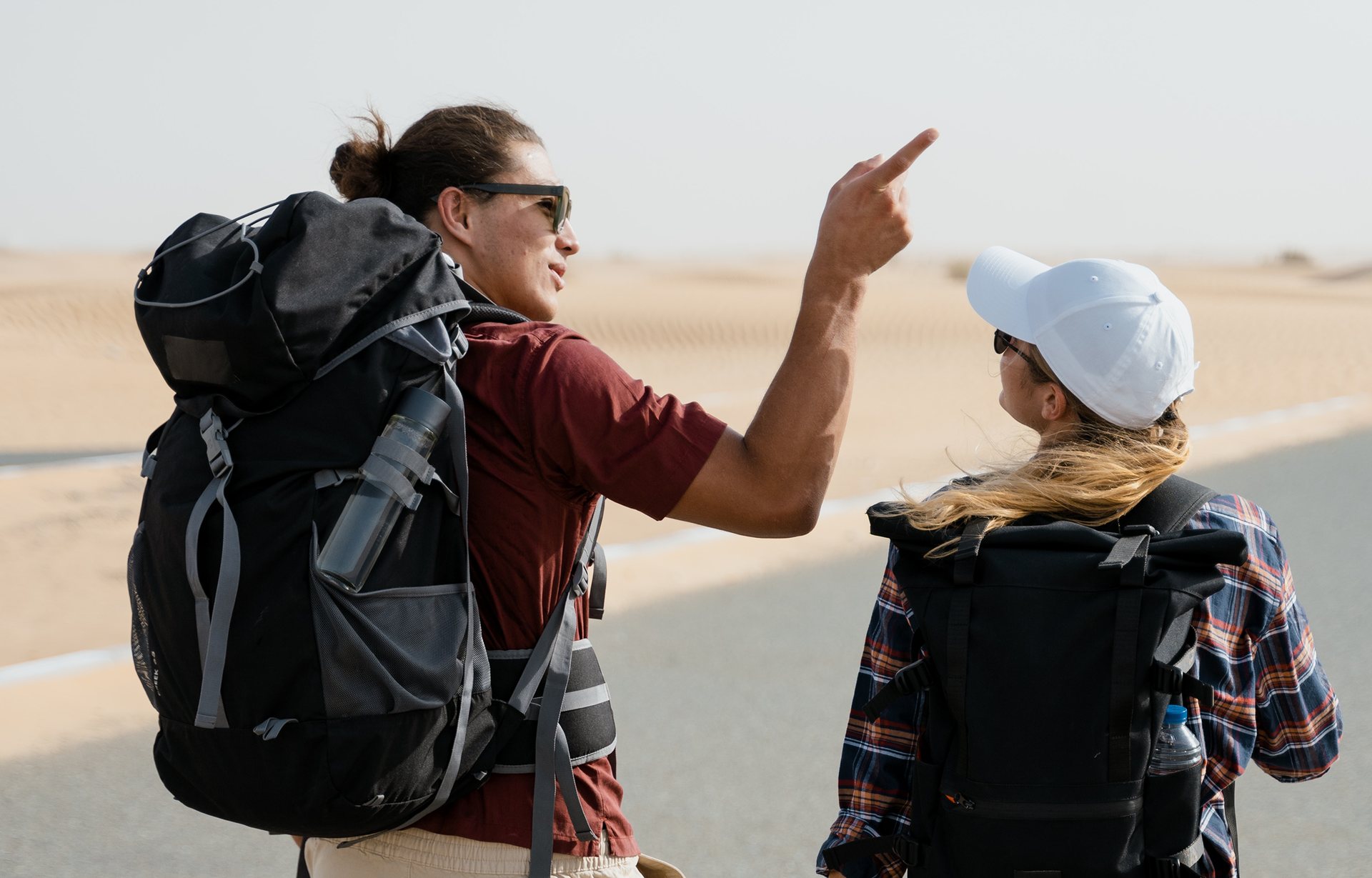 Zdjęcie przedstawia w ujęciu od tyłu parę młodych ludzi z plecakami, którzy idą drogą. Po lewej stronie widoczny jest chłopak, z długimi, ciemnymi włosami związanymi w kok, ubrany w brązową bluzkę z krótkim rękawem i z założonymi okularami słonecznymi. Na plecach ma duży, czarny plecak, do którego przymocowana jest butelka termiczna. Prawą rękę ma zgiętą w łokciu i podniesioną, a palcem wskazującym pokazuje na coś znajdującego się u góry po prawej stronie. Na prawo od niego widoczna jest dziewczyna, z długimi, jasnymi włosami, związanymi w kucyk i schowanymi pod białą czapką z daszkiem. Dziewczyna ubrana jest w koszulę w kratę, a na plecach ma czarny plecak. Głowę ma zwróconą w lewo i patrzy na chłopaka.