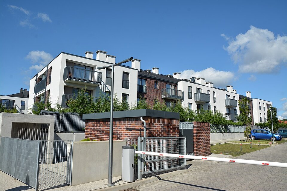 Na zdjęciu widoczne jest Osiedle Apartamenty Warszewo w Szczecinie. Wjazd na osiedle jest zabezpieczony szlabanem. Ulice osiedlowe są zbudowane z kostki brukowej. Po lewej stronie widoczny jest budynek mieszkalny - segmentowiec. Ma on dwie kondygnacje, płaski dach i białe oraz brązowe ściany. 