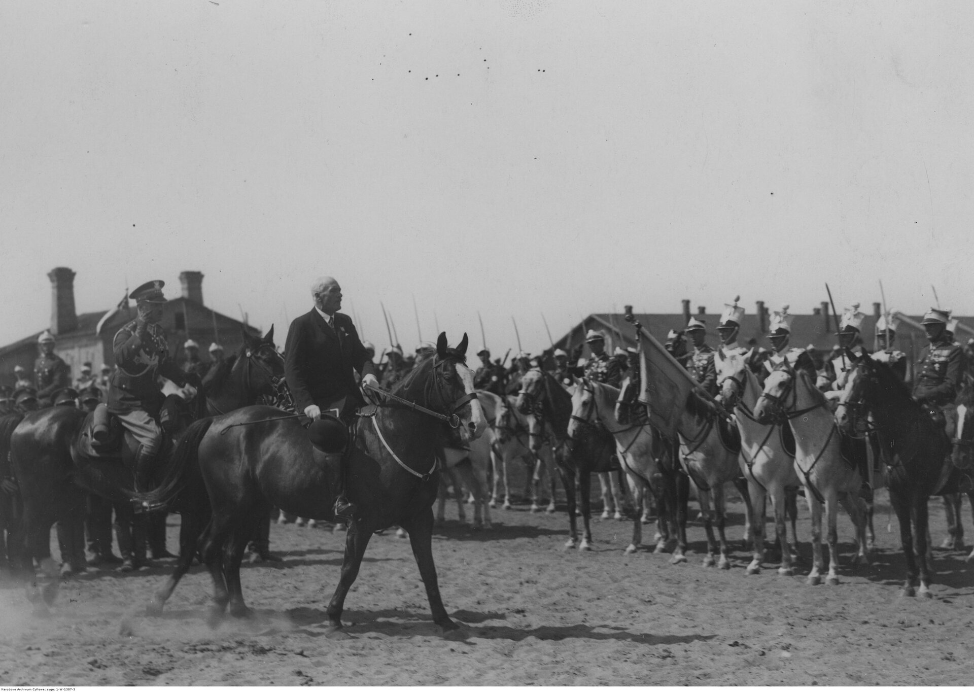 Zdjęcie przedstawia Ignacego Mościckiego, jadącego na koniu w trakcie uroczystości. Przed nim stoją mężczyźni w mundurach ze sztandarami, siedzący na koniach.