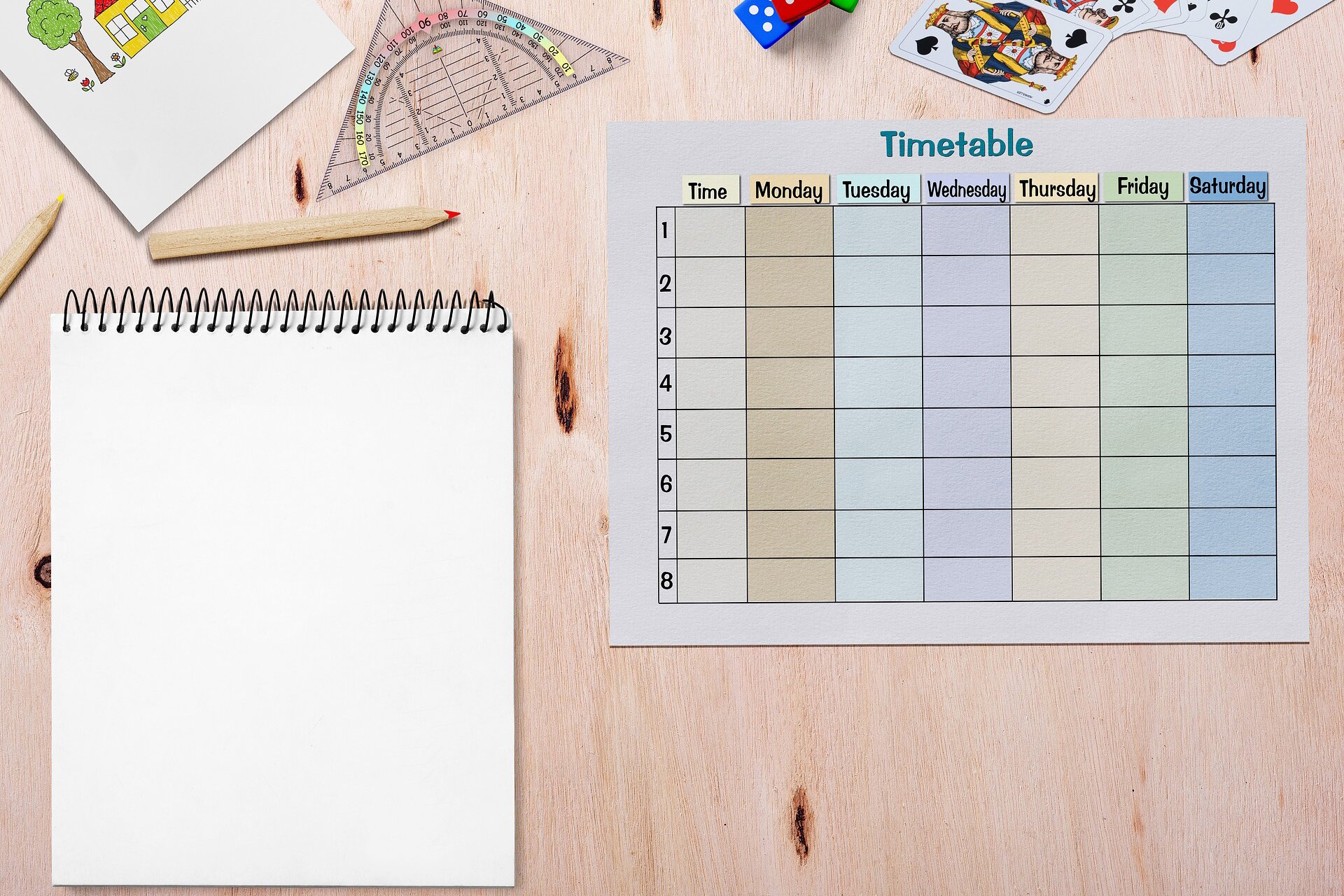 Ilustracja przedstawia widok na biurko z góry. U góry znajduje się rysunek, ekierka, kostki do gry, karty oraz dwie kredki. W głównej części ilustracji znajduje się notatnik z czystą, niezapisaną kartką oraz niewypełniony plan w formie tabeli (timetable) na każdy dzień tygodnia od poniedziałku do soboty włącznie, z miejscami na wpisanie zadań lub lekcji. 