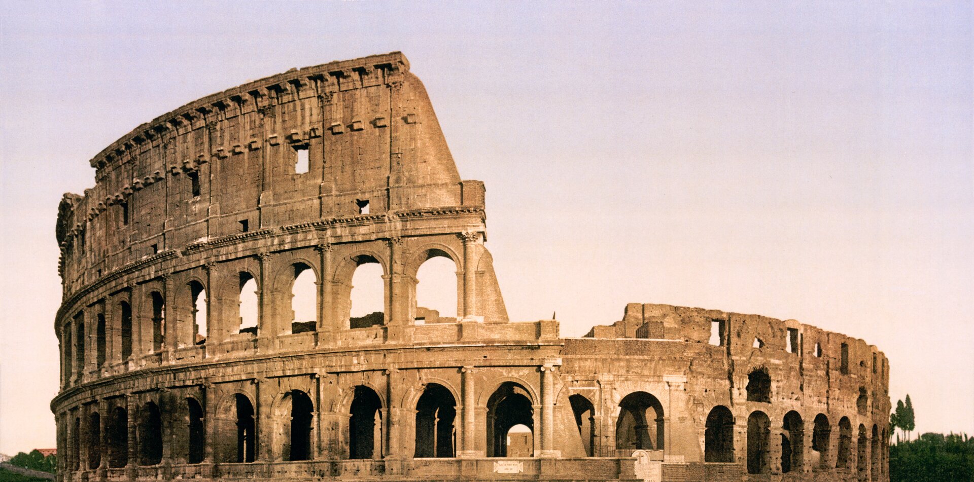 Widok współczesny na ruiny rzymskiego Koloseum. Ukazany w formie panoramicznej, widoczne zniszczenia górnej partii arkad od połowy zdjęcia w stronę prawej krawędzi fotografii. W dolnej części fotografii widoczne arkady z kolumnadą w proporcjonalnych odstępach. Obiekt sfotografowany za dnia przy wykorzystaniu filtru fotograficznego.