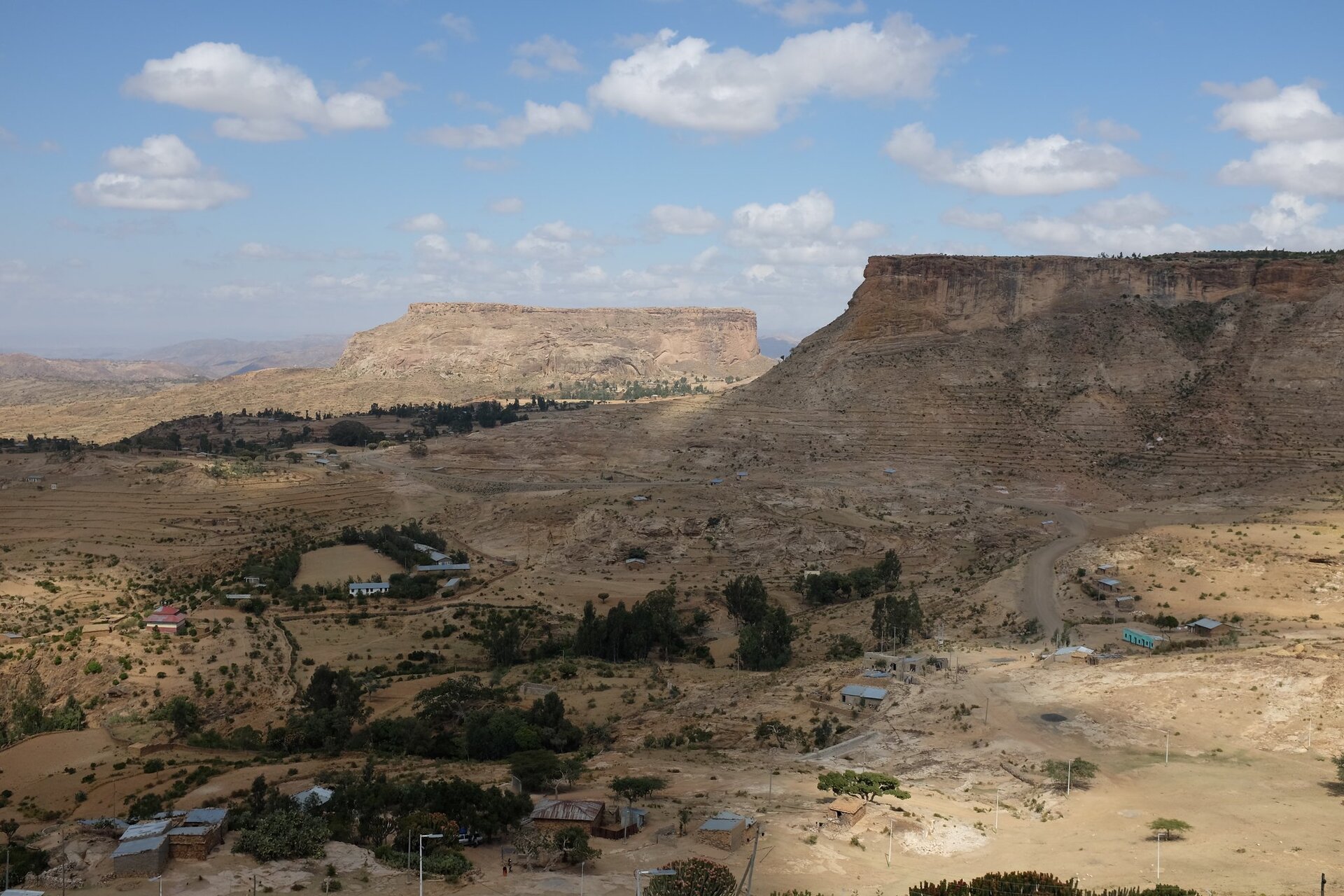 Fotografia przedstawiająca rozległy, półpustynny teren. Roślinność ograniczona, gdzieniegdzie jedynie niewielkie krzewy lub drzewa, poza tym jałowa gleba. Co kilkadziesiąt metrów pojedyncze, niewielkie zabudowania. Na drugim planie wysokie wzniesienia.