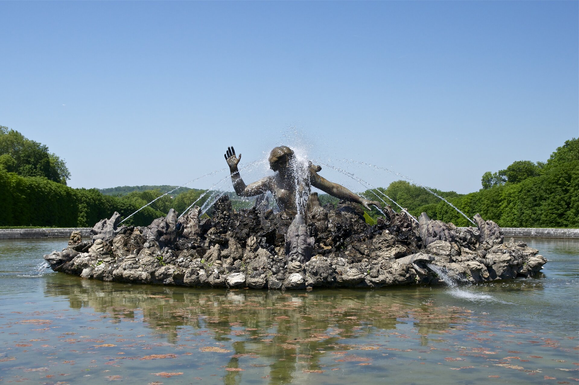 Zdjęcie przedstawia fontannę. W jej centrum znajduje się, widoczna do pasa, sylwetka nagiej kobiety z długimi, upiętymi włosami. Dookoła rozmieszczono skały, spomiędzy których wystają głowy bestii o wielkich zębach. Z ich paszczy tryskają strumienie wody. W oddali widać drzewa.