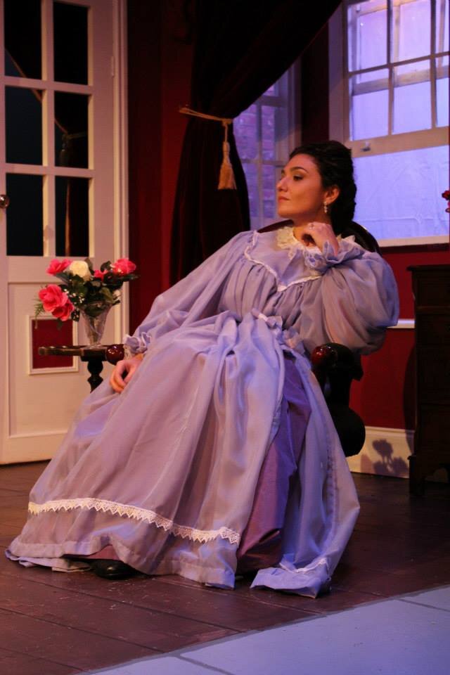 Fotografia przedstawia młodą kobietę siedzącą na fotelu. Kobieta ma ciemne włosy upięte w kok. Lekko unosi głowę. Patrzy na coś z wysoka.  Siedzi wygodnie, pełna dystyngowanej swobody. Prawą rękę opuściła wzdłuż ciała, lewą wspiera na poręczy fotela. Kobieta ubrana jest w długą suknię koloru fioletowego. Obok fotela mały stolik, a na nim wazon z kwiatami. Za kobietą okno. Po lewej stronie drzwi i ozdobna bordowa zasłona.