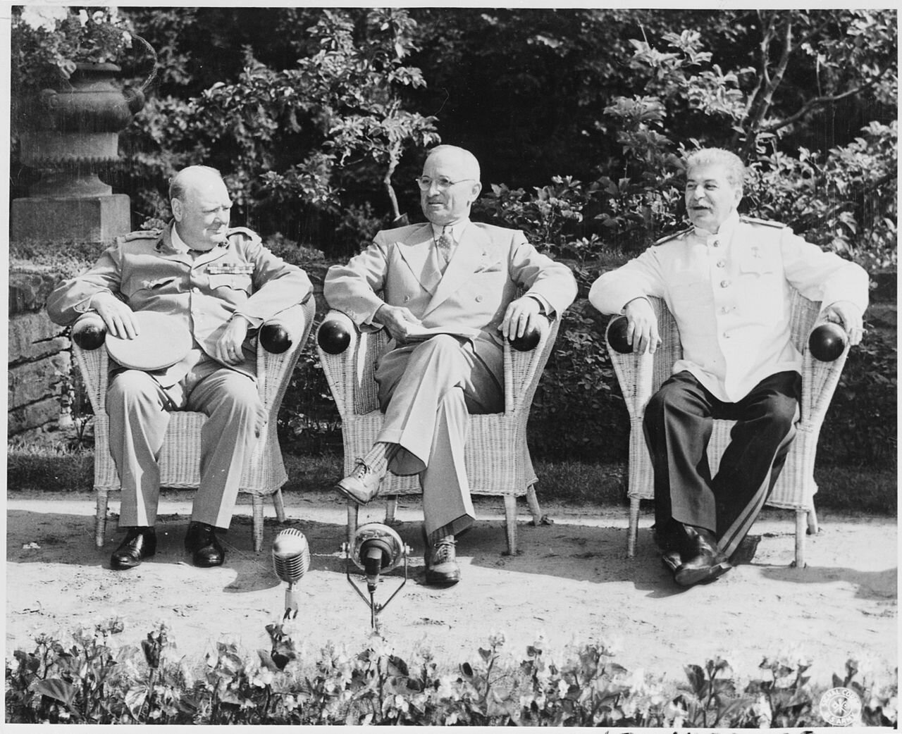 Zdjęcie przedstawia trzech starszych mężczyzn, którzy siedzą na krzesłach. Mężczyźni po lewej i prawej stronie są ubrani w mundury. Mężczyzna na środku ubrany jest w garnitur i nosi okulary. Mężczyzna po lewej jest łysiejący, w ręku trzyma kapelusz. Mężczyzna po prawej ma krótkie włosy i gęste wąsy.