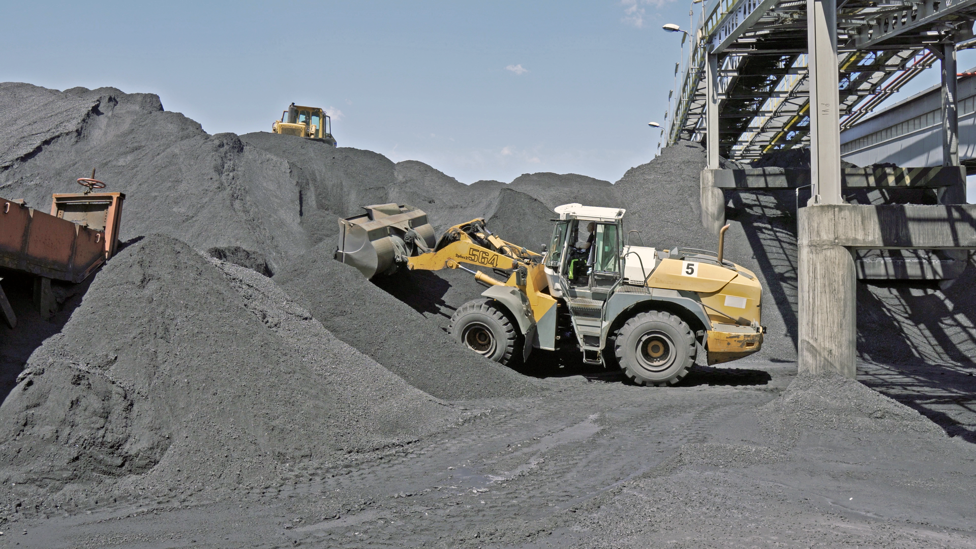 Na zdjęciu widać nagarnianie miału na składowisku. W centralnej części zdjęcia znajduje się duża czarna hałda miału. Po lewej stronie widoczny jest przenośnik taśmowy transportujący miał na hałdę. Po prawej stronie żółta ładowarka nagarnia miał na hałdę.