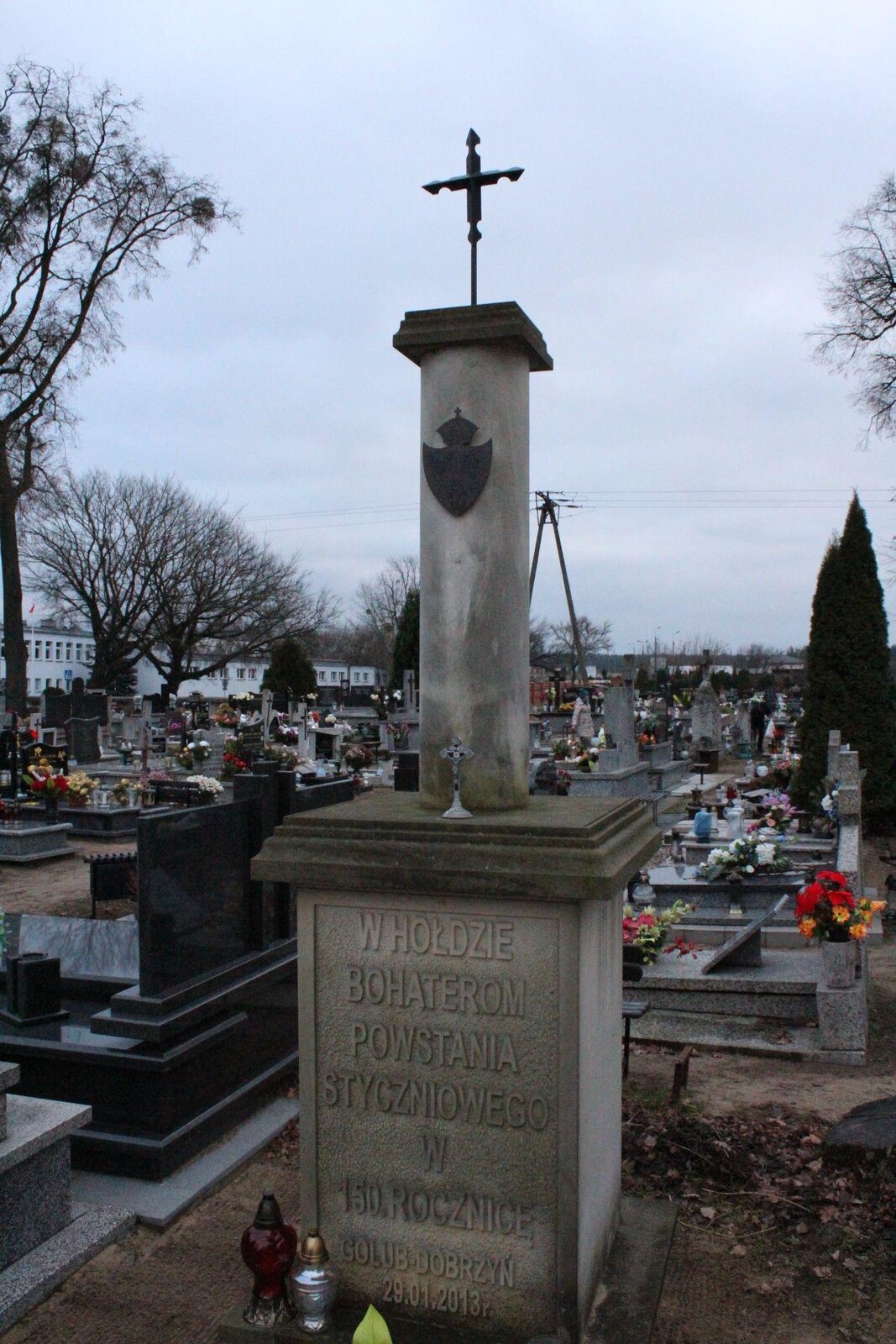 Zdjęcie przedstawia pomnik i betonową kolumnę, u szczytu której znajduje się metalowy krzyż. Poniżej, do kolumny, został przytwierdzony metalowy herb. Kolumna stoi na graniastosłupie, na którym wyryto napis: "W hołdzie bohaterom powstania styczniowego w sto pięćdziesiątą rocznicę. Gołub Dobrzyń, 29.01.2013".  W tle widoczne są współczesne nagrobki, a także bezlistne drzewa. Niebo jest błękitne, bezchmurne. Zdjęcie zostało wykonane w świetle dziennym.