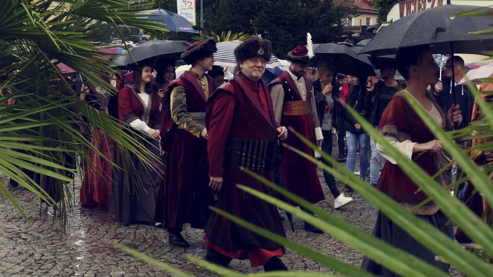Na zdjęciu znajduje się grupa ludzi idących ulicą. Mężczyźni ubrani są w długie bordowe płaszcze. Kobiety noszą długie suknie. Wszyscy mają na głowach czapki obszyte futrem. W tle zdjęcia znajdują się ludzie oraz budynki.