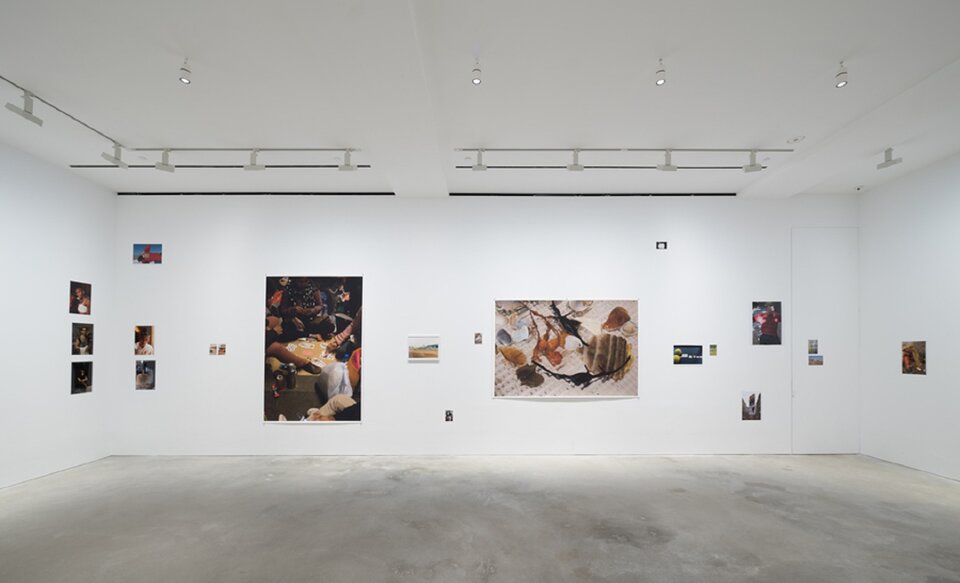 Ilustracja przedstawia fragment wystawy Wolfganga Tillmansa. Ukazuje ona białe wnętrze z działami sztuki na ścianach. Wiszą obrazy oraz fotografie, przedstawiające osoby lub krajobrazy.