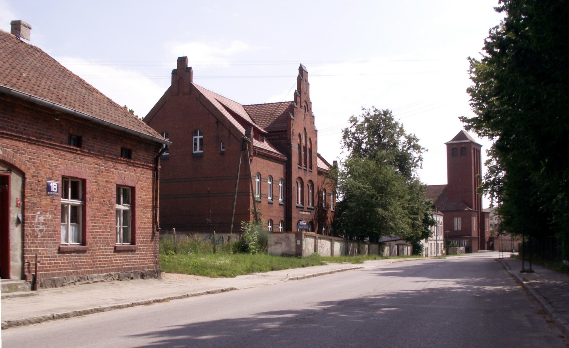 Zdjęcie przedstawia niewielkie budynki wykonane z czerwonej cegły. Są prostej konstrukcji, posiadają okna o białych ramach. Lekko podniszczone. Stoją wzdłuż asfaltowej drogi na terenie miasteczka.