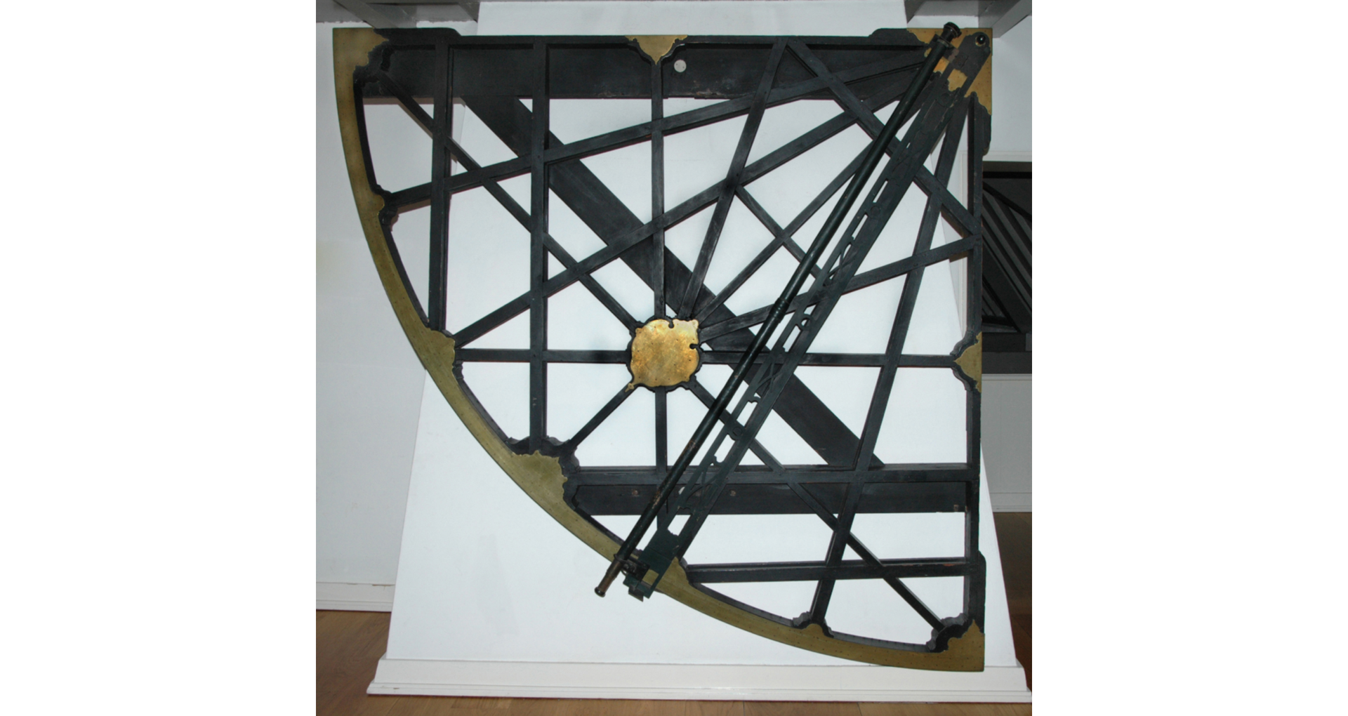 Zdjęcie przedstawia kwadrant, czyli przyrząd służący do pomiaru kątów. Jego kształt przypomina ćwiartkę okręgu, w którego wnętrzu znajduje się wiele przecinających się ze sobą odcinków. Posiada również element ruchomy, który pozwala wyznaczyć kąt.