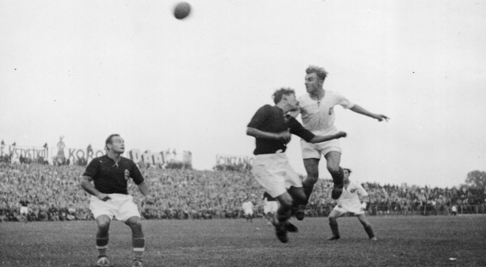 Zdjęcie przedstawia scenę z boiska do gry w piłkę nożną. Widocznych jest dwóch piłkarzy w wyskoku do piłki. Są to piłkarze z przeciwnych drużyn. Jeden z mężczyzn ubrany jest w białe spodenki i czarną koszulkę, a drogi w białe spodenki i białą koszulkę. W tle widoczne są trybuny, na których zgromadzili się licznie widzowie. 