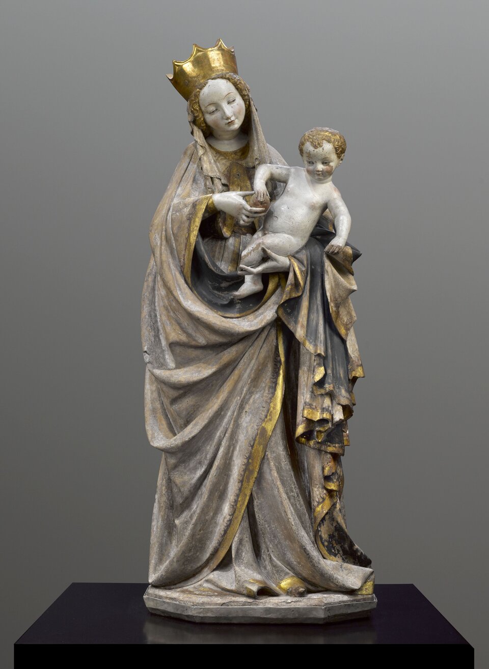 Ilustracja przedstawia rzeźbę Madonny trzymającej dziecko na rękach. Madonna jest ubrana w złotą szatę oraz ma koronę na głowie. Dziecko, które jest trzymane przez kobietę jest gołe oraz ma rude włosy.