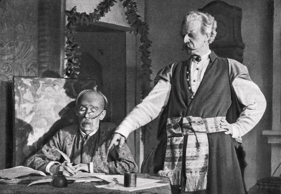 Fotografia przedstawiająca starszych mężczyzn znajdujących się wewnątrz pomieszczenia. Jeden z nich siedzi przy stole. Ma owalną twarz, jest łysy, ma cienkie brwi, prosty nos i sumiaste wąsy. Nosi okulary, pochyla się nad kartką papieru. W prawej dłoni trzyma pióro. Notuje coś. Na stole leżą kartki papieru i butelka z atramentem. Mężczyzna jest ubrany w koszulę i surdut. Obok stoi mężczyzna ubrany w żupan i kontusz. Jego talia jest przewiązana szerokim pasem słuckim ozdobionym poziomymi paskami i frędzlami przyszytymi do krótszych boków. Postać ma owalną twarz, zakola, gęste, lekko falujące, sterczące włosy. Brwi mężczyzny są zmarszczone, oczy głęboko osadzone, a nos haczykowaty. Ma również sumiaste wąsy. Prawą rękę wysuwa do przodu i palcem wskazuje list. Lewą rękę opiera o biodro.