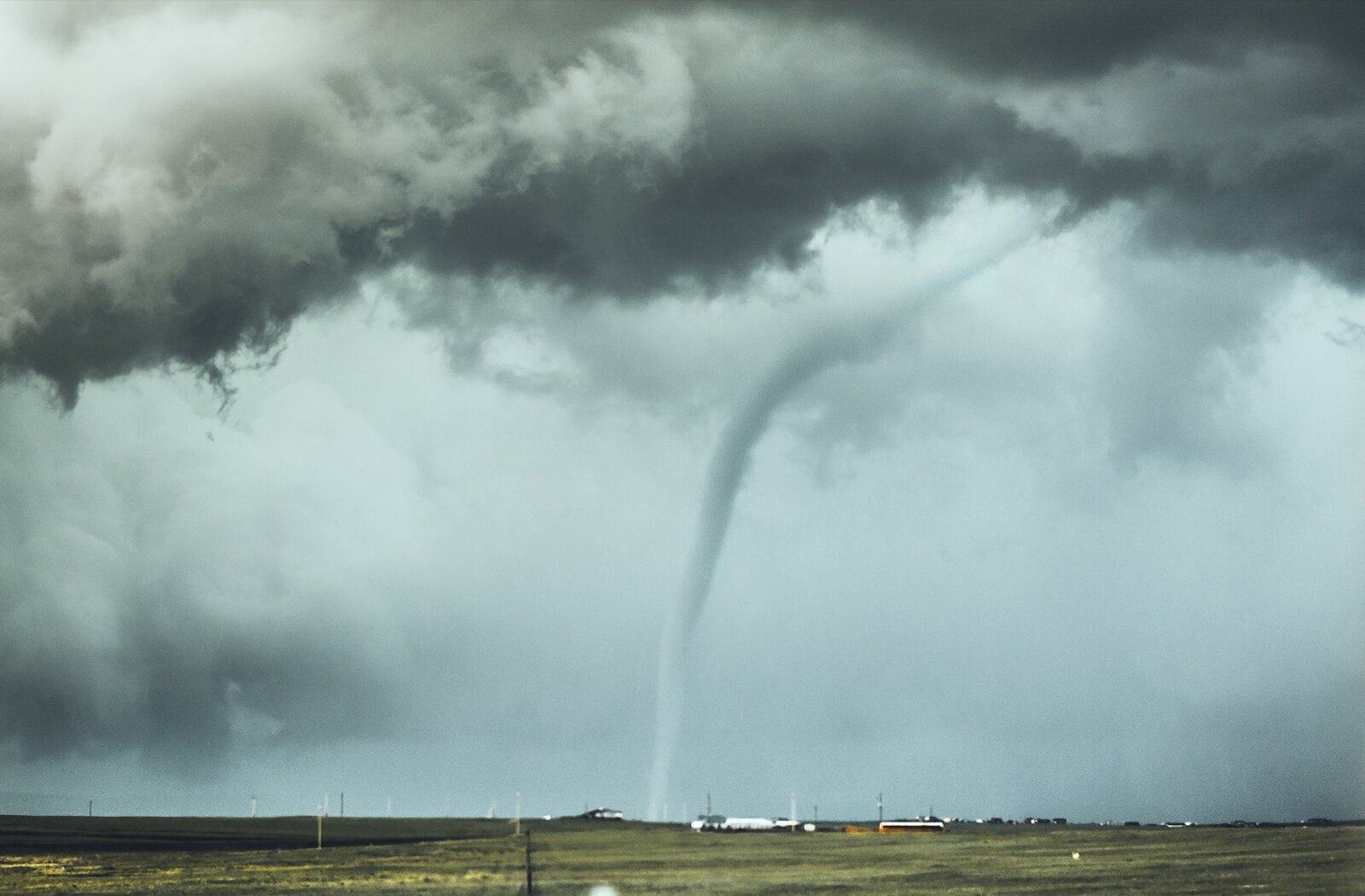 Zdjęcie przedstawia trąbę powietrzną znajdująca się nad polem. Trąba ma kolor biało‑szary. W miejscu, w którym znajduje się trąba powietrza, widoczne są  pojedyncze zabudowania. Niebo jest siwo‑czarne, z głębiącymi się stalowymi chmurami. 