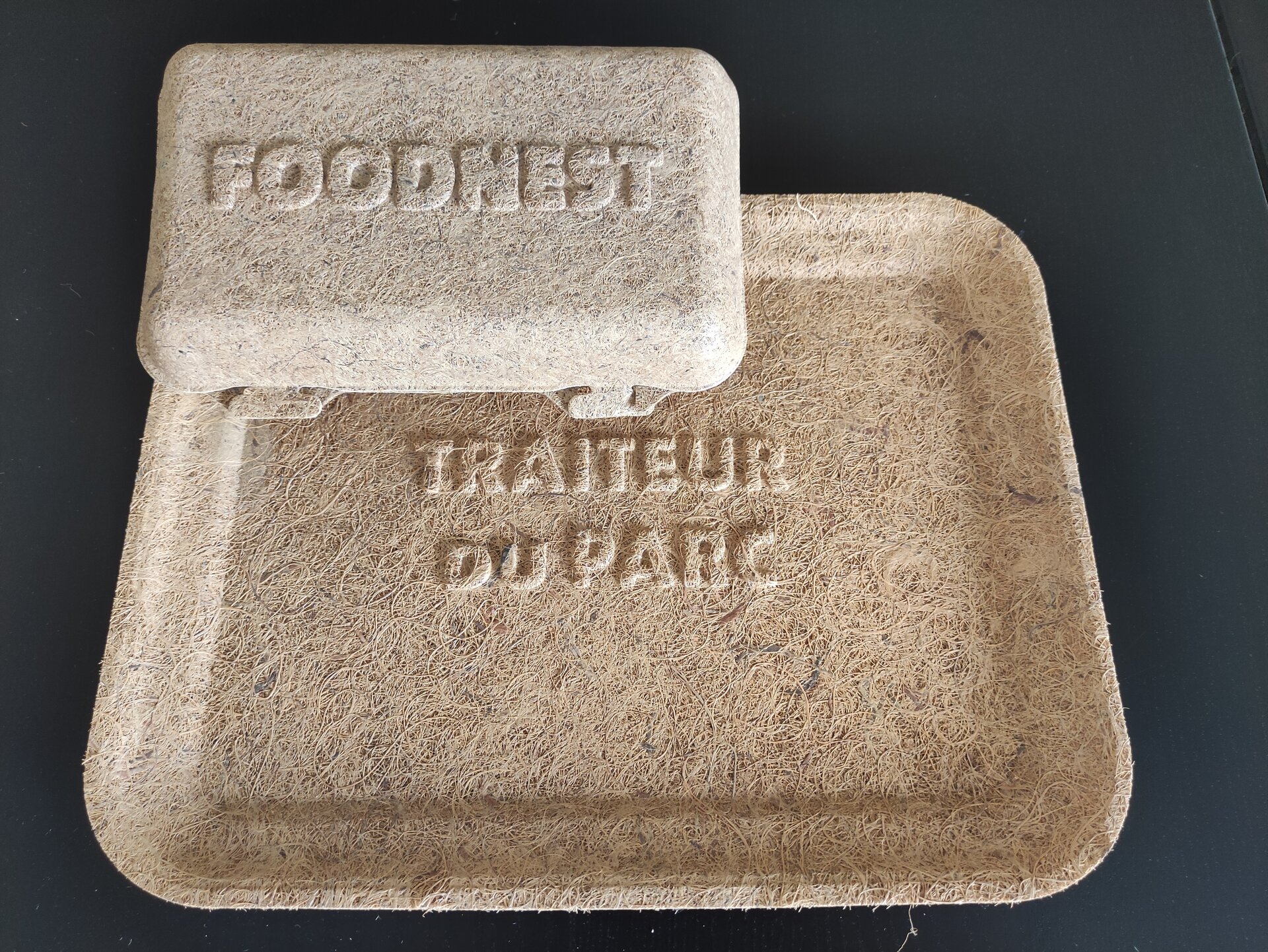 Na zdjęciu widoczne dwa płaskie opakowania. Pierwsze przypomina tackę, drugie płaską pokrywkę. Oba są prostokątne i mają wytłoczone napisy na wierzchu.