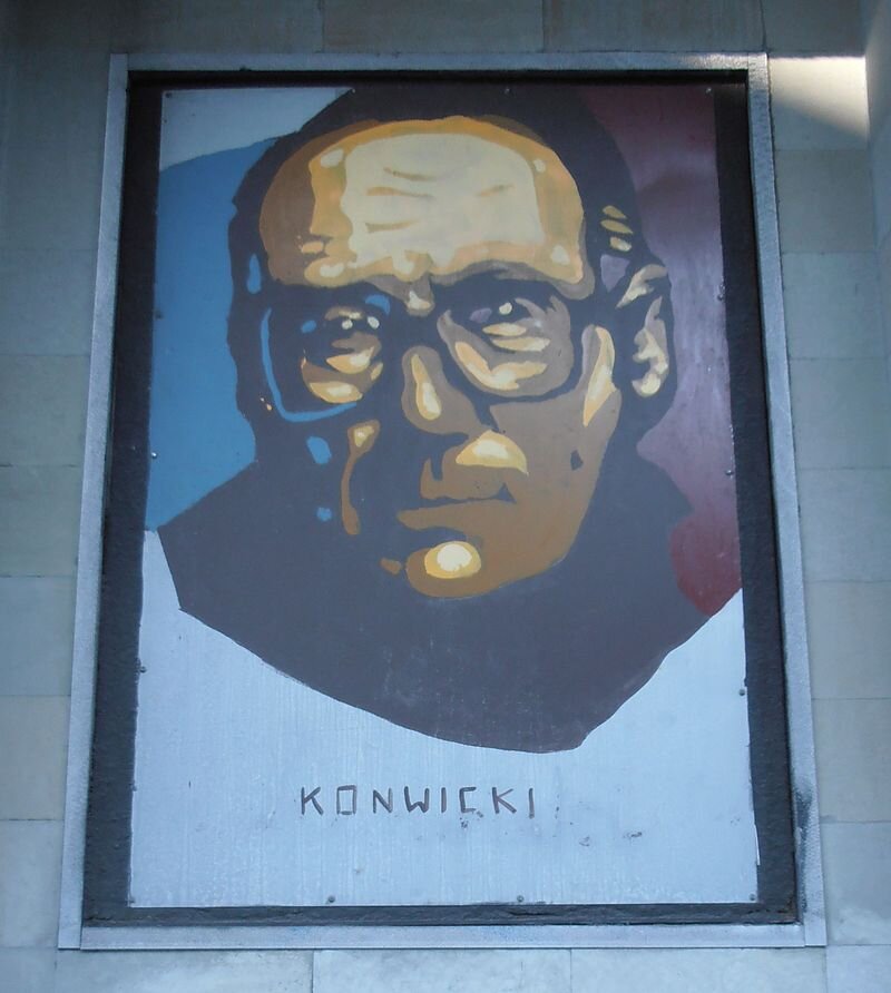 Zdjęcie przedstawia tablicę z wizerunkiem starszego mężczyzny. Mężczyzna jest łysiejący, ma zmarszczki na twarzy oraz okulary w grubych oprawkach. Pod spodem jest napis: Konwicki.