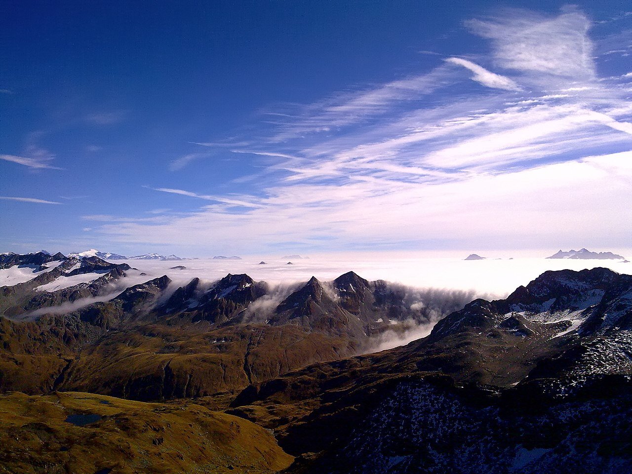 Zdjęcie przedstawia pasmo górskie. Po prawej stronie zdjęcia ostre szczyty wyłaniają się spod warstwy chmur. Warstwa chmur zasłania dolinę. Na niebie białe chmury przybierają kształt pasów.