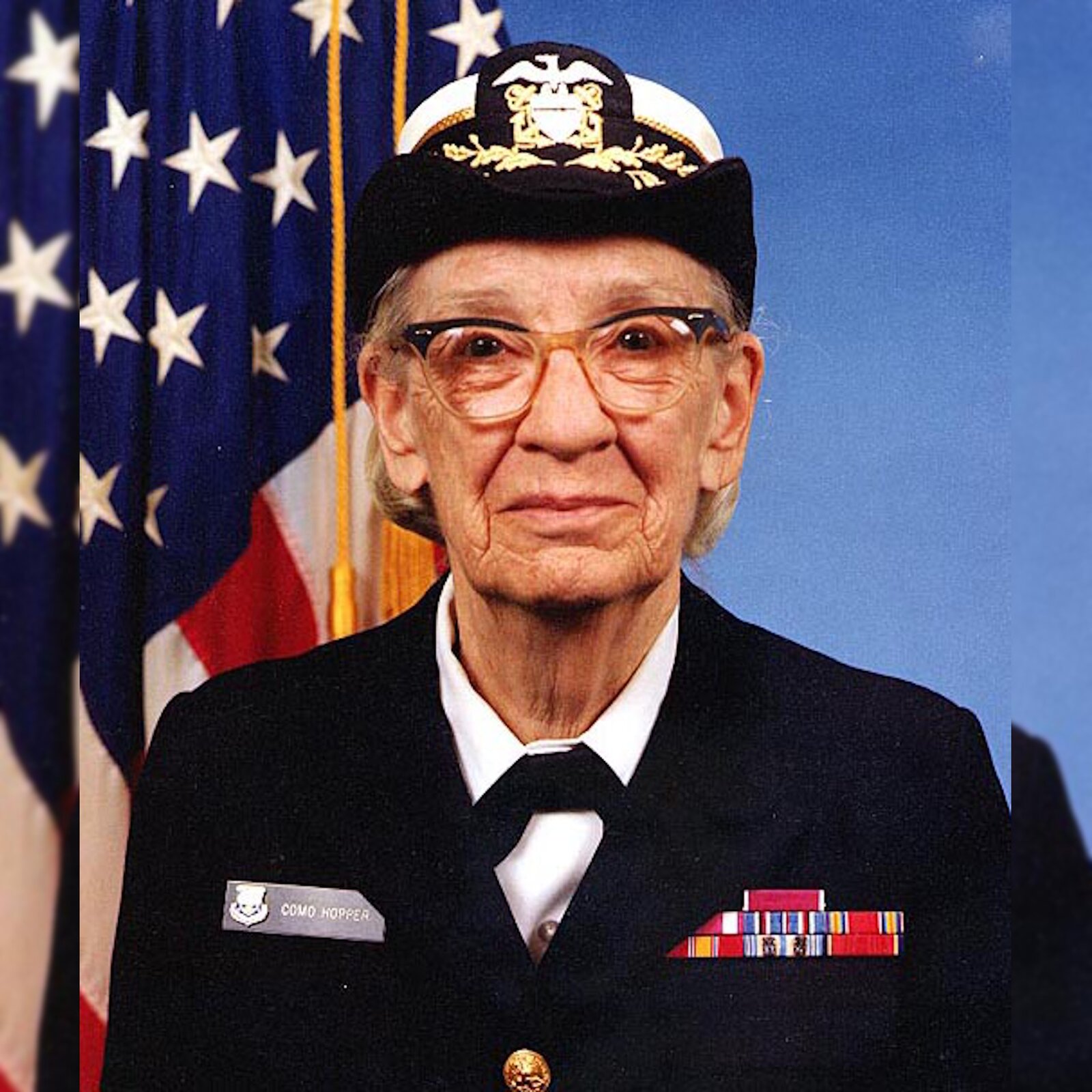 Zdjęcie przedstawia kobietę w podeszłym wieku. Kobieta ma wydatne uszy i nos, na nosie okulary. Ubrana jest w mundur. Na prawej klapie marynarki znajduje się naszywka z tekstem: komo hoper. Kobieta ma na głowie granatowo‑białą czapkę. W tle widać fragment flagi amerykańskiej.