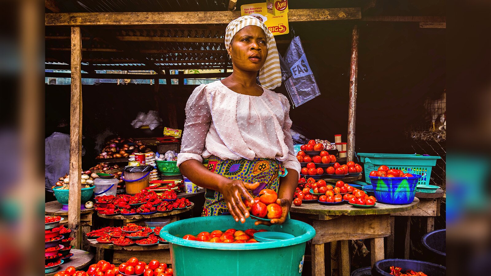 Zdjęcie przedstawia czarnoskórą kobietę sprzedającą pomidory. Kobieta jest ubrana w różową bluzkę i kolorową spódnicę, a na głowie ma przewiązaną chustę w biało-żółte paski. W lewej dłoni trzyma talerzyk z umytymi pomidorami. Prawą dłoń ma mokrą i dotyka nią pomidorów na talerzyku. Poniżej jej dłoni widoczna jest duża zielono-niebieska misa, w której pływają w wodzie pomidory oraz dwa małe talerzyki. W tle za nią widoczny jest stragan z ustawionymi w różnych pojemnikach pomidorami oraz czerwoną papryką. W głębi widoczne są pojemniki z czosnkiem i cebulą oraz dwa plastikowe wiaderka. U góry widoczna jest żółta etykieta z napisem w języku angielskim: Your Assurance for great Meals.