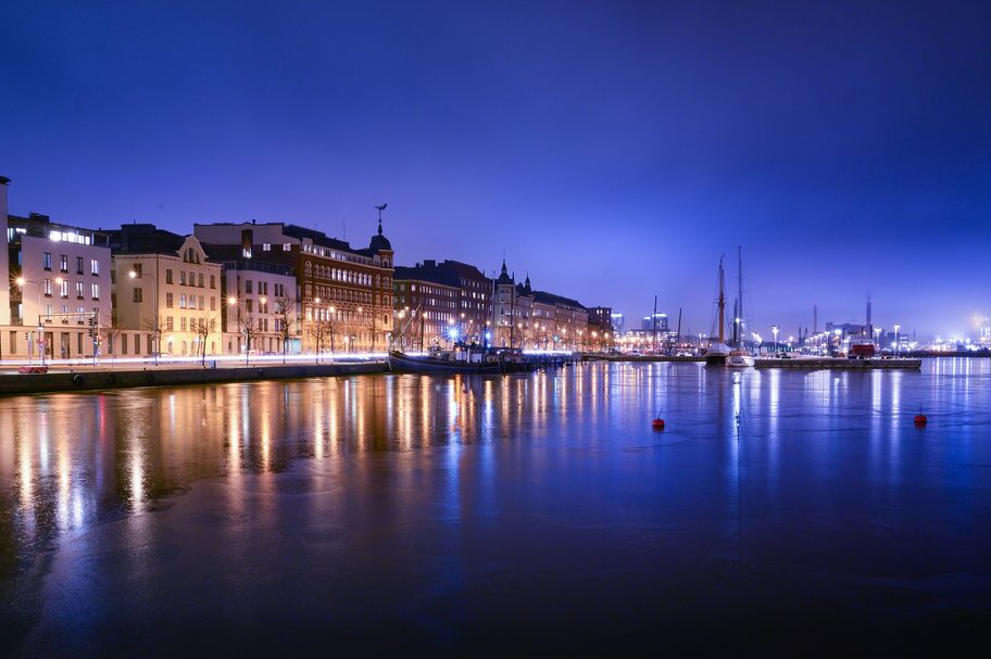 Zdjęcie przedstawia widok na Helsinki. Budynki znajdują się nad wodą, w tle port i liczne statki.