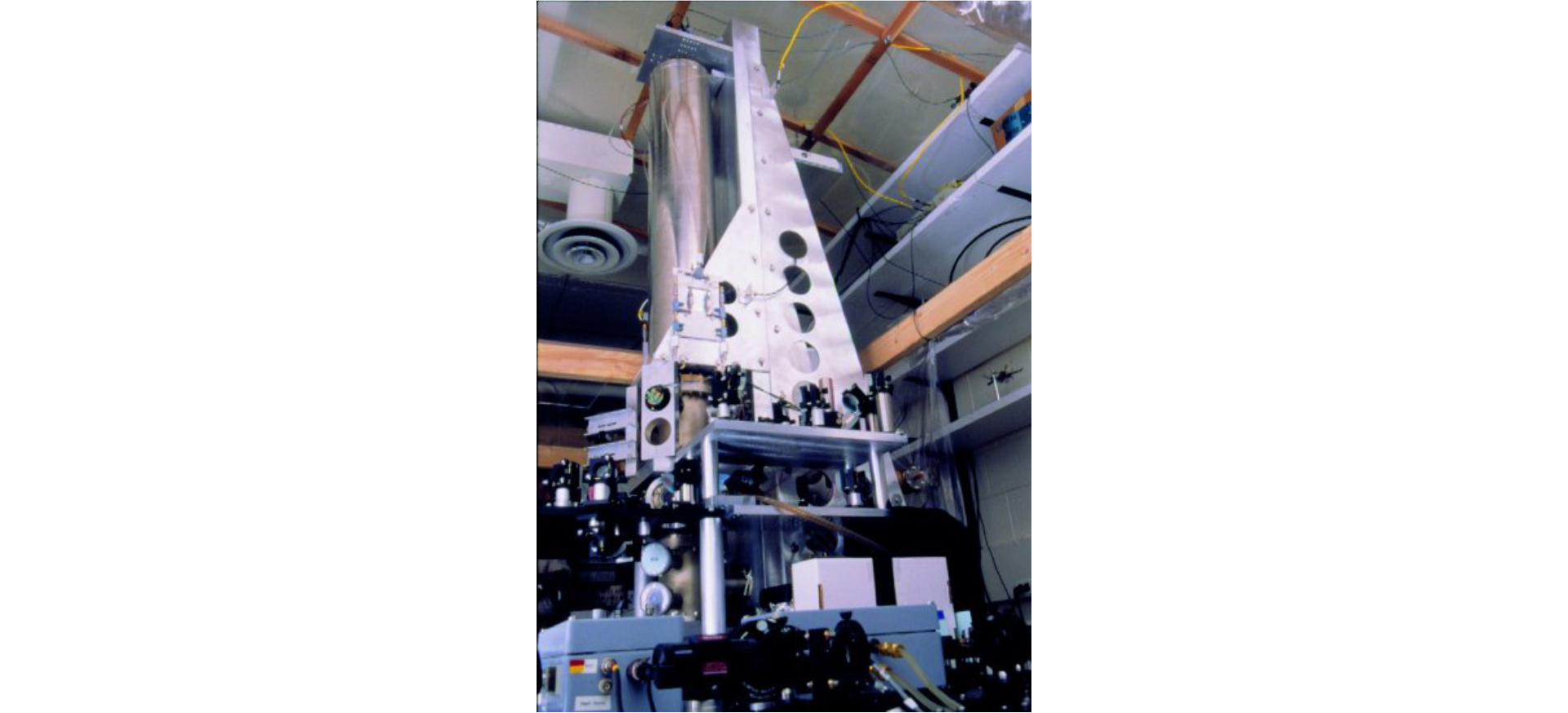 Zdjęcie przedstawia dużą, skomplikowaną konstrukcję, sięgającą sufitu pomieszczenia. Jej największym elementem jest gruba, metalowa rura. Otaczają ją mniejsze urządzenia na stelażach oraz skrzynki z podłączoną elektroniką.