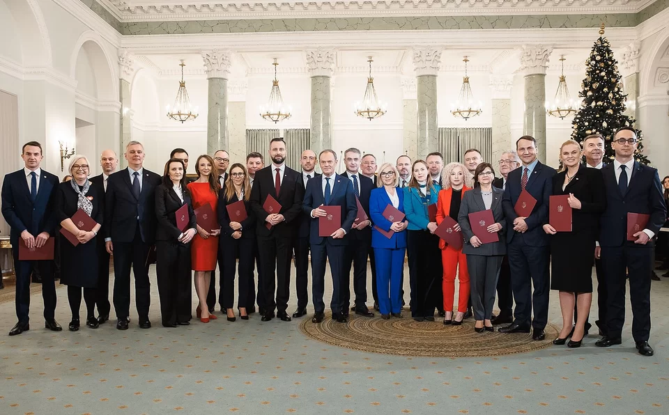 Jasna, reprezentacyjna sala Pałacu Prezydenckiego z marmurowymi kolumnami i żyrandolami. Na pierwszym planie 27 osób w strojach formalnych, większość trzyma bordowe teczki z godłem. W centrum, nieco wysunięta do przodu, stoi osoba pełniąca funkcję premiera; po bokach ministrowie i ministrynie. Po prawej widoczna wysoka, przystrojona choinka. Wszyscy patrzą w obiektyw; atmosfera uroczysta, zdjęcie po ceremonii powołania rządu.