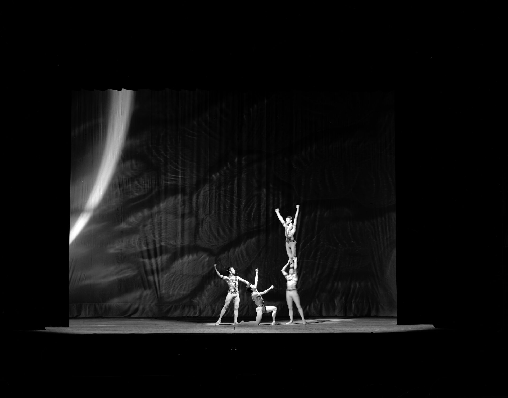 Czarno-białe zdjęcie spektaklu baletowego wykonane przez fotografa André Cros. Na zdjęciu widoczna jest scena, na której czterej mężczyźni w obcisłych strojach wykonują akrobacje. Po prawej stronie sceny stoi mężczyzna, na którego ramionach stoi kolejny z rękoma wzniesionymi ku górze. Tworzą pewnego rodzaju wieżę. Dwaj pozostali znajdują się po lewej stronie sceny, jeden stoi, drugi klęczy. Obaj wznoszą ręce w kierunku mężczyzny znajdującego się na górze. 